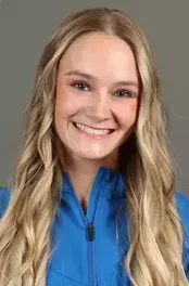 Smiling woman with long blonde hair wearing a blue jacket against a plain gray background.