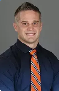 Portrait of a man with short hair wearing a dark shirt and orange striped tie, smiling against a neutral background.