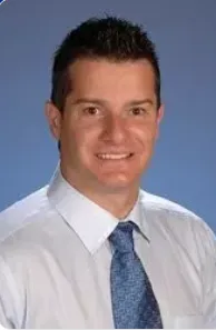 Smiling man with short dark hair wearing a white shirt and blue patterned tie, against a blue background.
