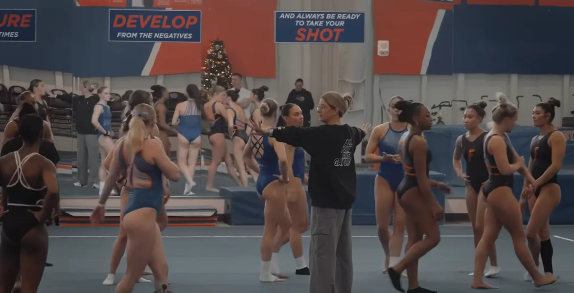 Male gymnast in a red top and white pants performing a hold on gymnastic rings in a bright gymnasium.