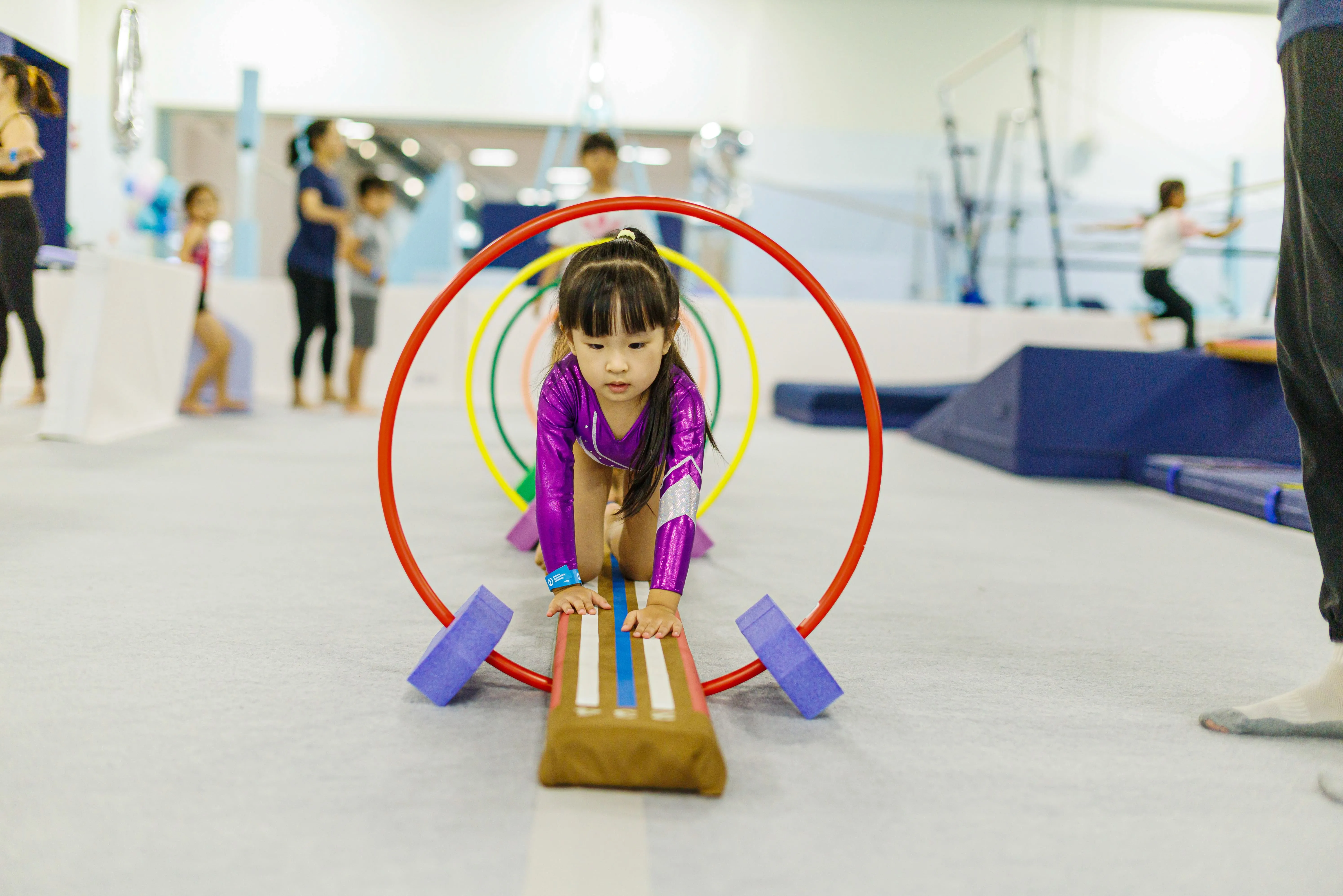 Children sitting on mats in a gym performing stretching exercises with arms raised.