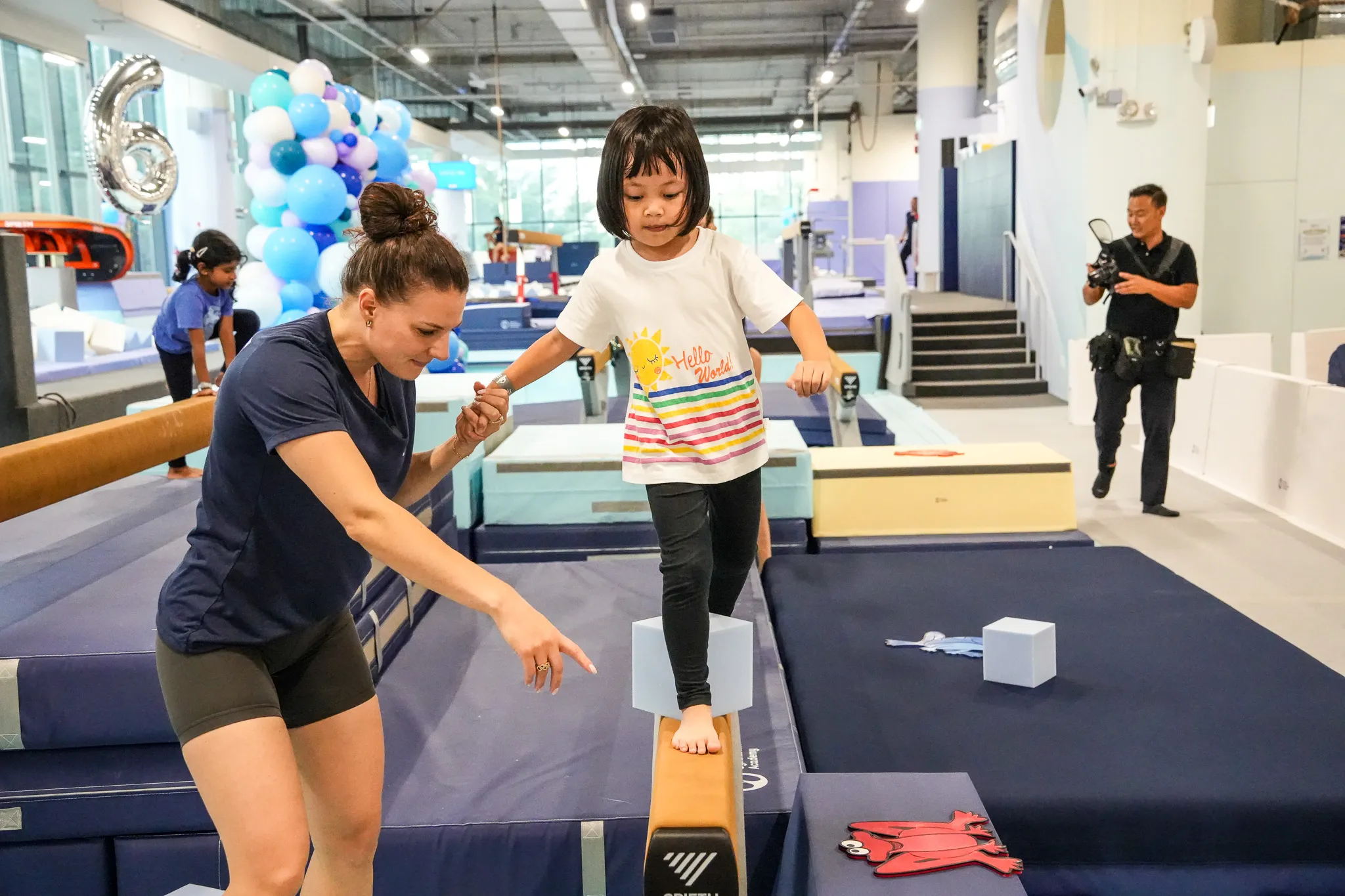 A young girl in a blue leotard practicing gymnastics with the support of a coach kneeling behind her in a gym.