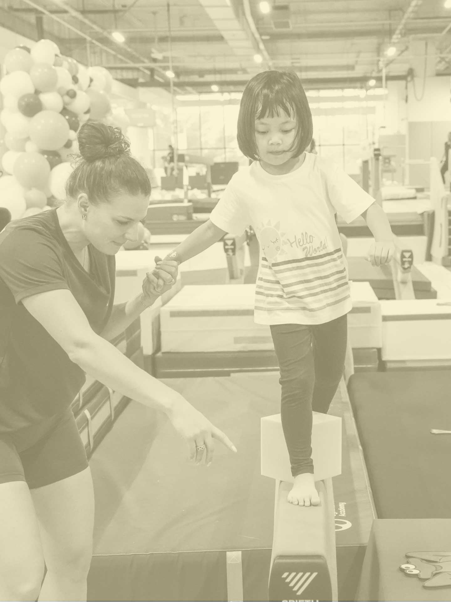 A coach assisting a young girl in gymnastics, holding her hand as she prepares to perform near a balance beam.