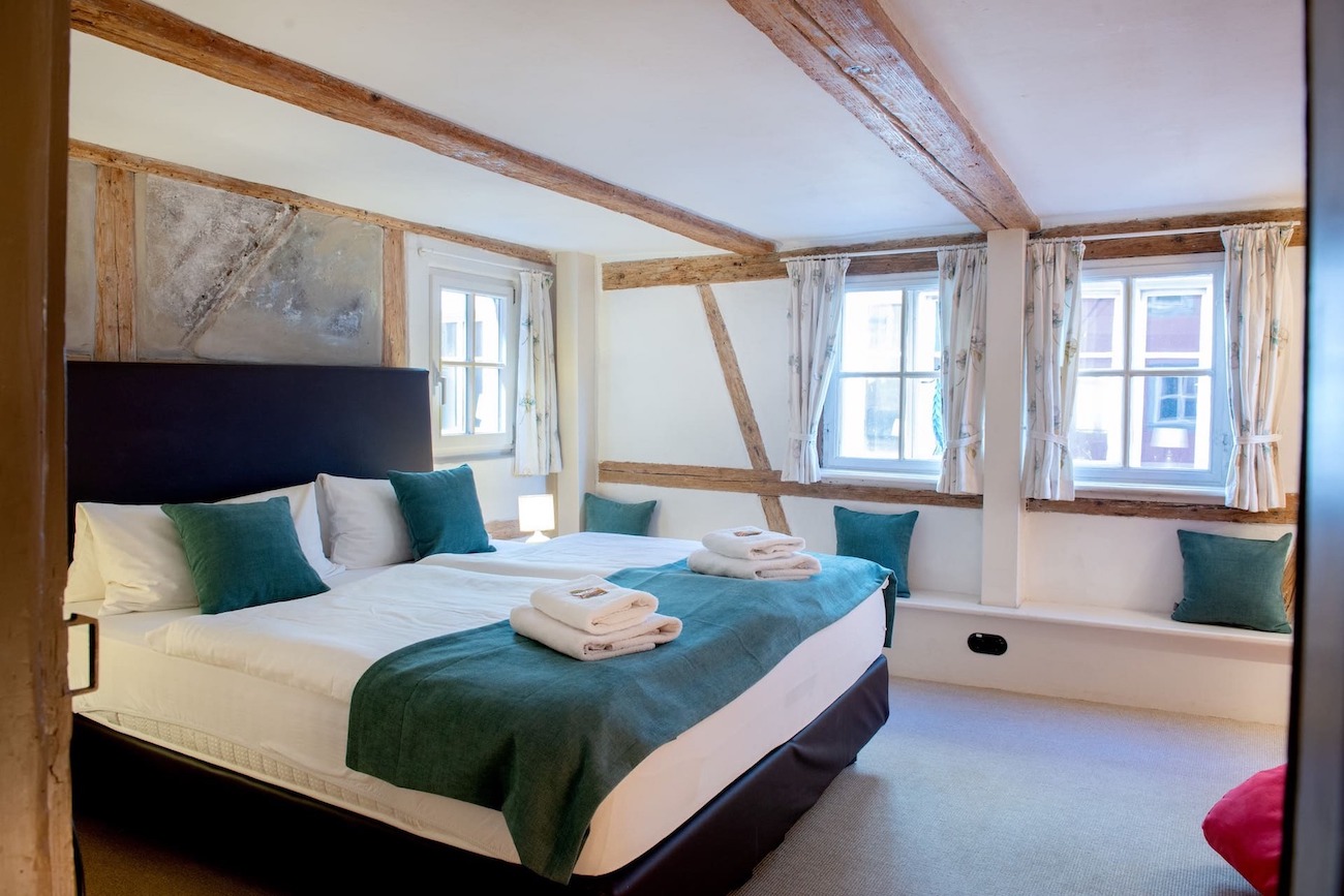 Schlafzimmer mit Kingsizebett, weißen Kopfkissen und Bettdecken, grüneren Dekokissen und Bettlaeufer, Sitzbank mit grünen Dekokissen, Fenster mit Vorhaengen