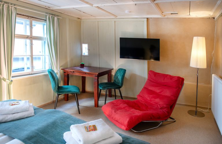 Wohnraum mit Tisch, 2 Stuehlen, Fernseher, Loungesessel, Fenster mit Vorhaengen