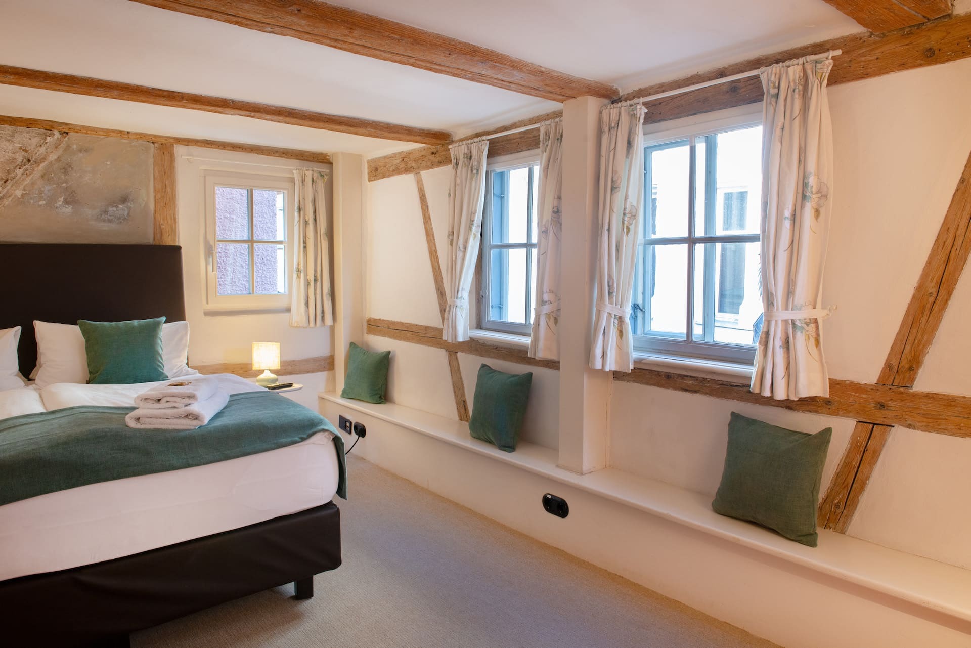 Schlafraum mit Kingsizebett und weißen Kissen und Bettdecken, grüne Dekokissen und Bettlaeufer, Fenster mit Vorhang, Nachttisch mit Nachttischlapme, Sitzbank mit Dekokissen