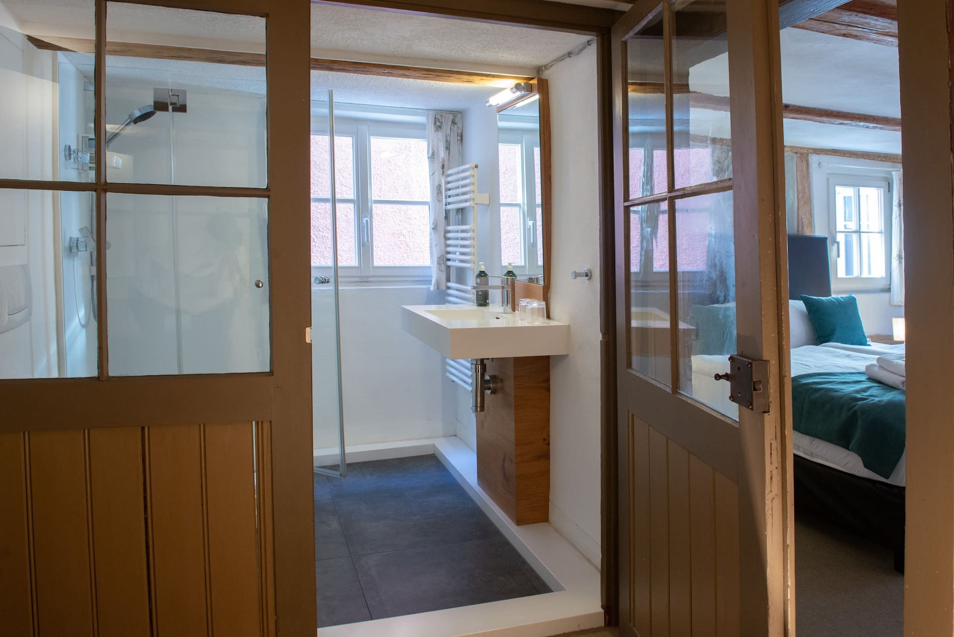 Blick in das Badezimmer mit Fenster, Waschbecken, Spiegel, Dusche, und in das Schlafzimmer mit Kingsizebett