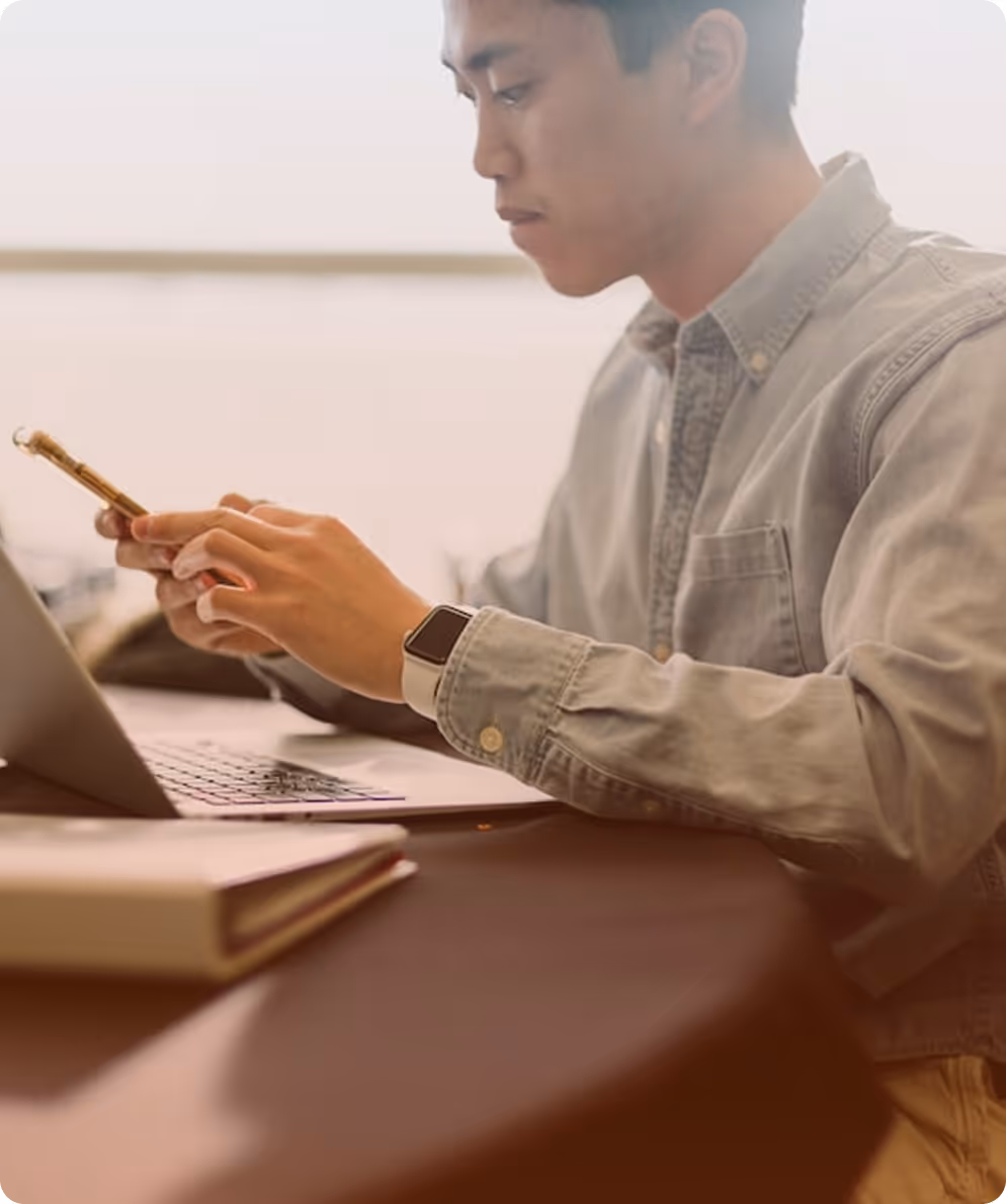 Man in a denim shirt uses smartphone and laptop at a table with a closed book nearby.
