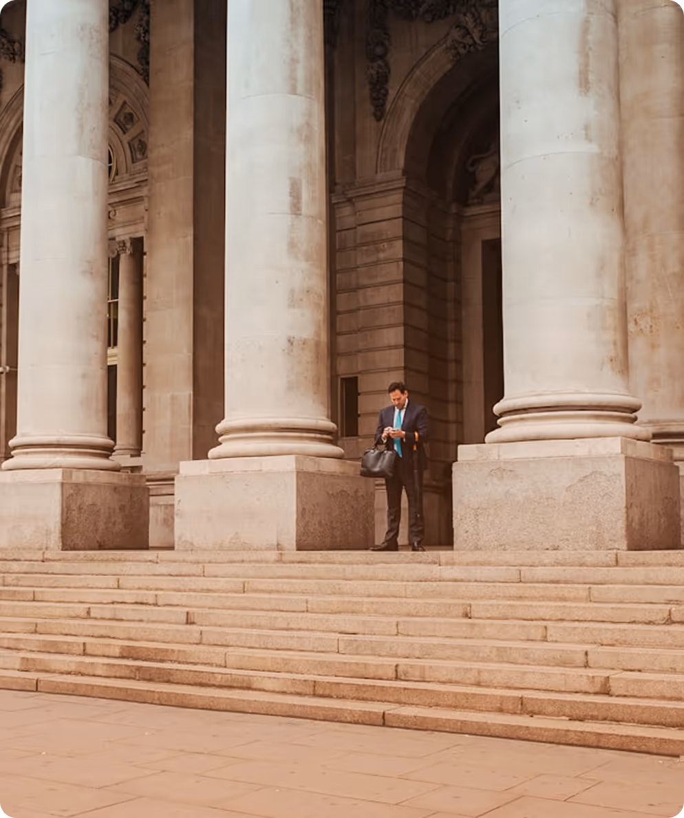 Man in suit holding bag and looking at phone standing on stone steps by large building columns.