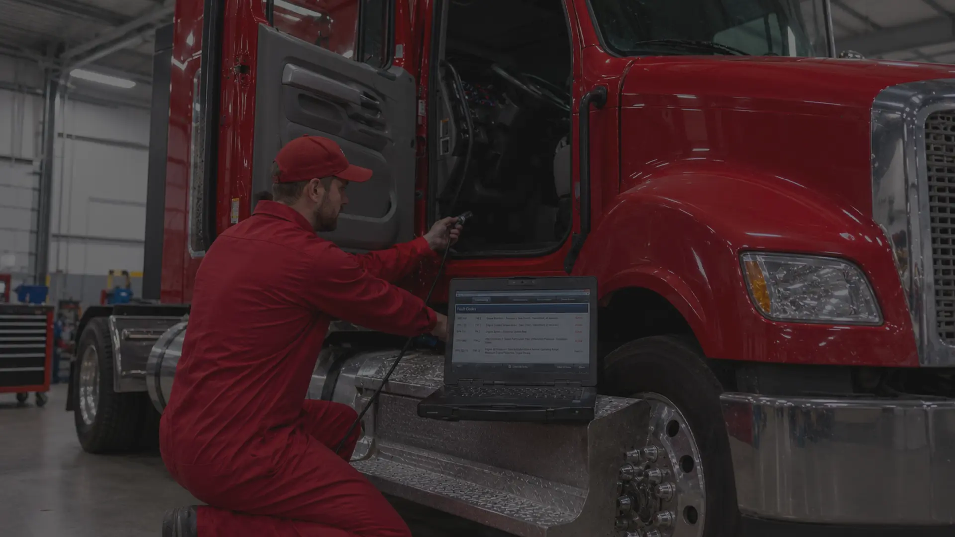 Fleet technician connecting a diagnostic laptop to the OBD port of a Class 8 semi truck inside a commercial repair shop, fault codes visible on screen