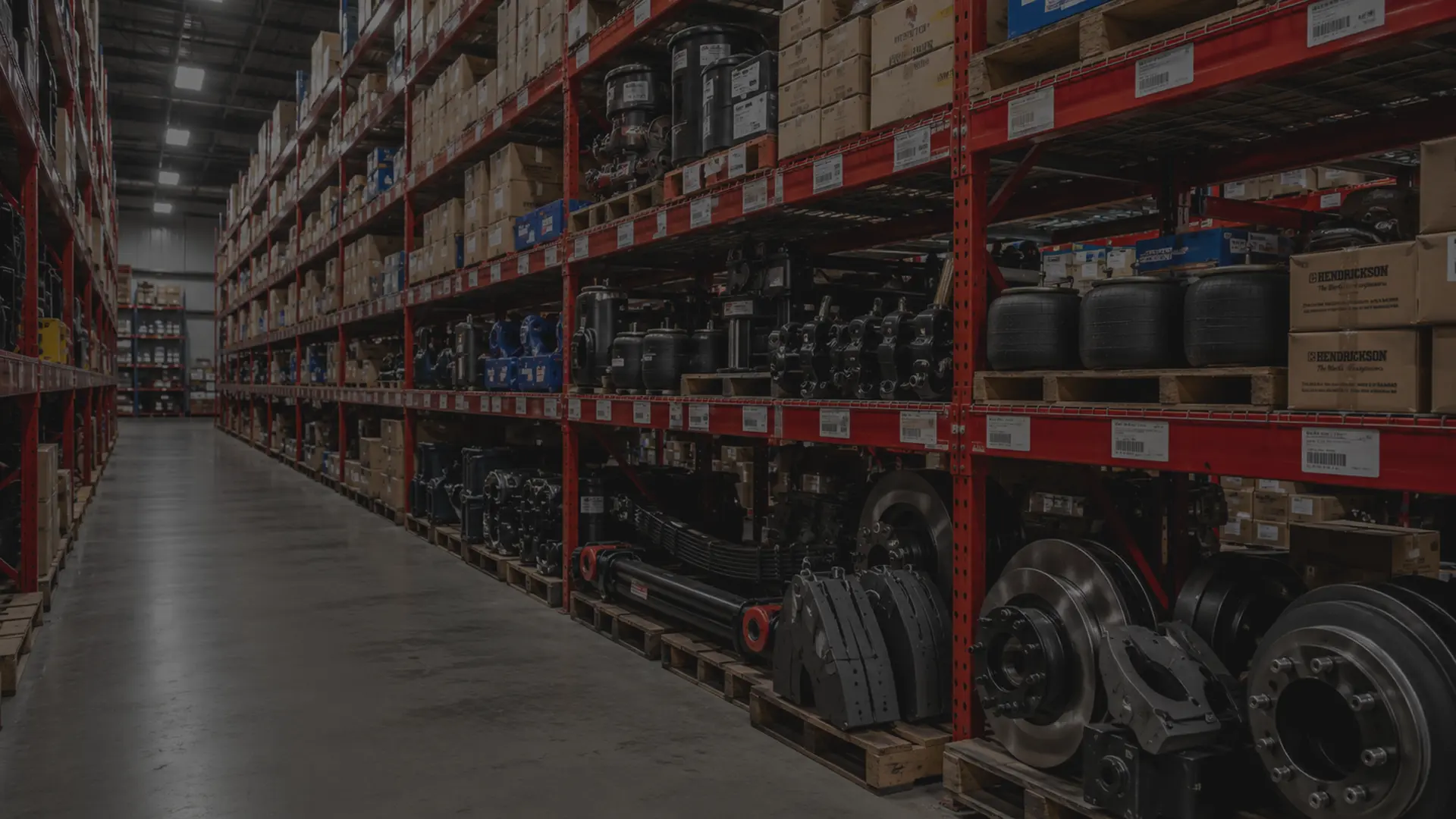 Rows of OEM and aftermarket commercial truck parts on shelving in a large fleet parts warehouse, brake components and suspension parts visible in foreground