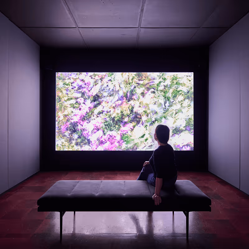 Person sitting on a bench in a dim gallery room, viewing a large abstract artwork with purple and green tones on the wall.