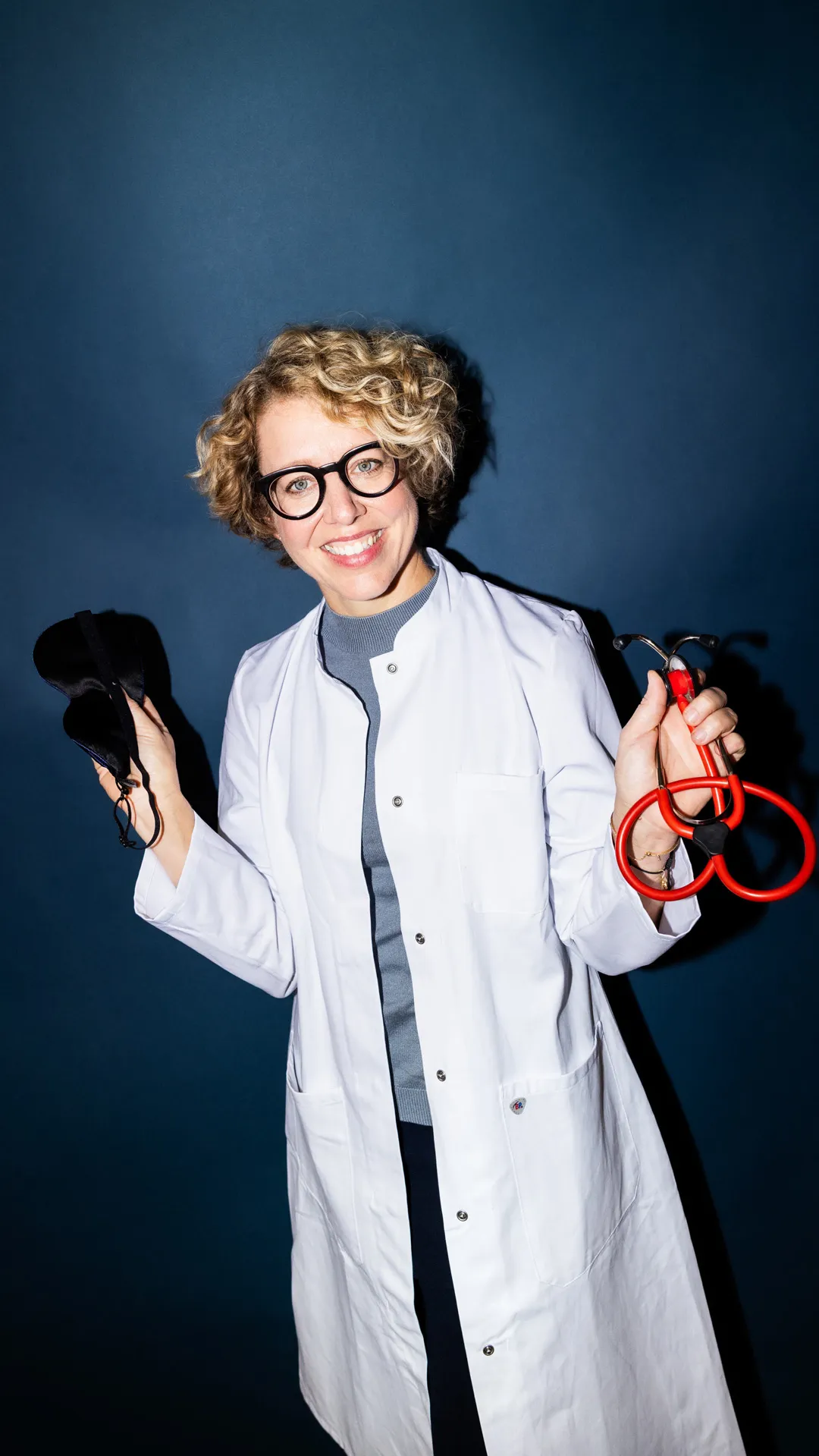 Lächelnde Ärztin mit kurzen lockigen Haaren, die eine schwarze Schutzmaske und ein rotes Stethoskop hält.