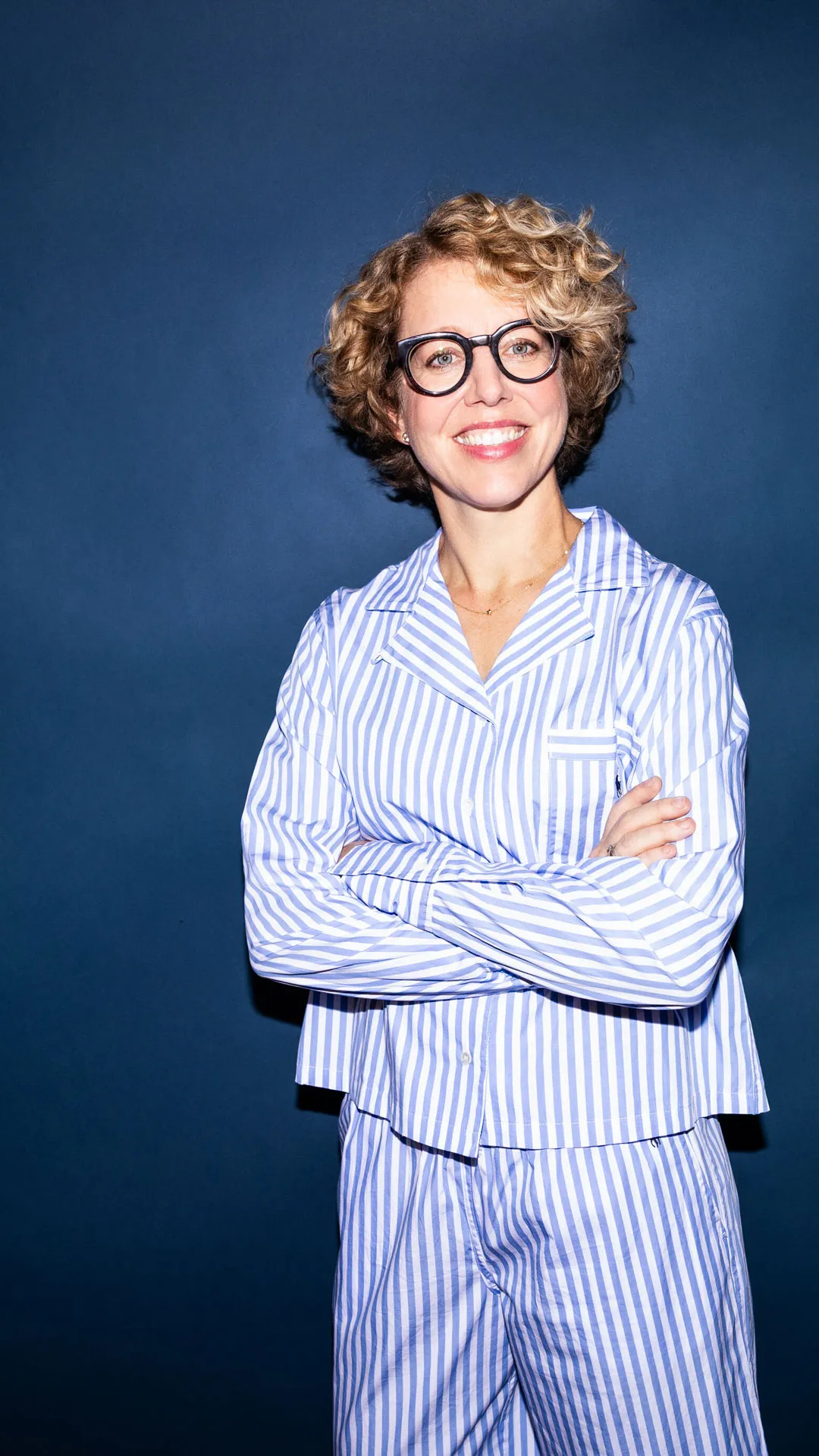 Frau mit lockigem kurzen blonden Haar und Brille trägt gestreifte blaue und weiße Pyjamahose und Hemd vor dunkelblauem Hintergrund.