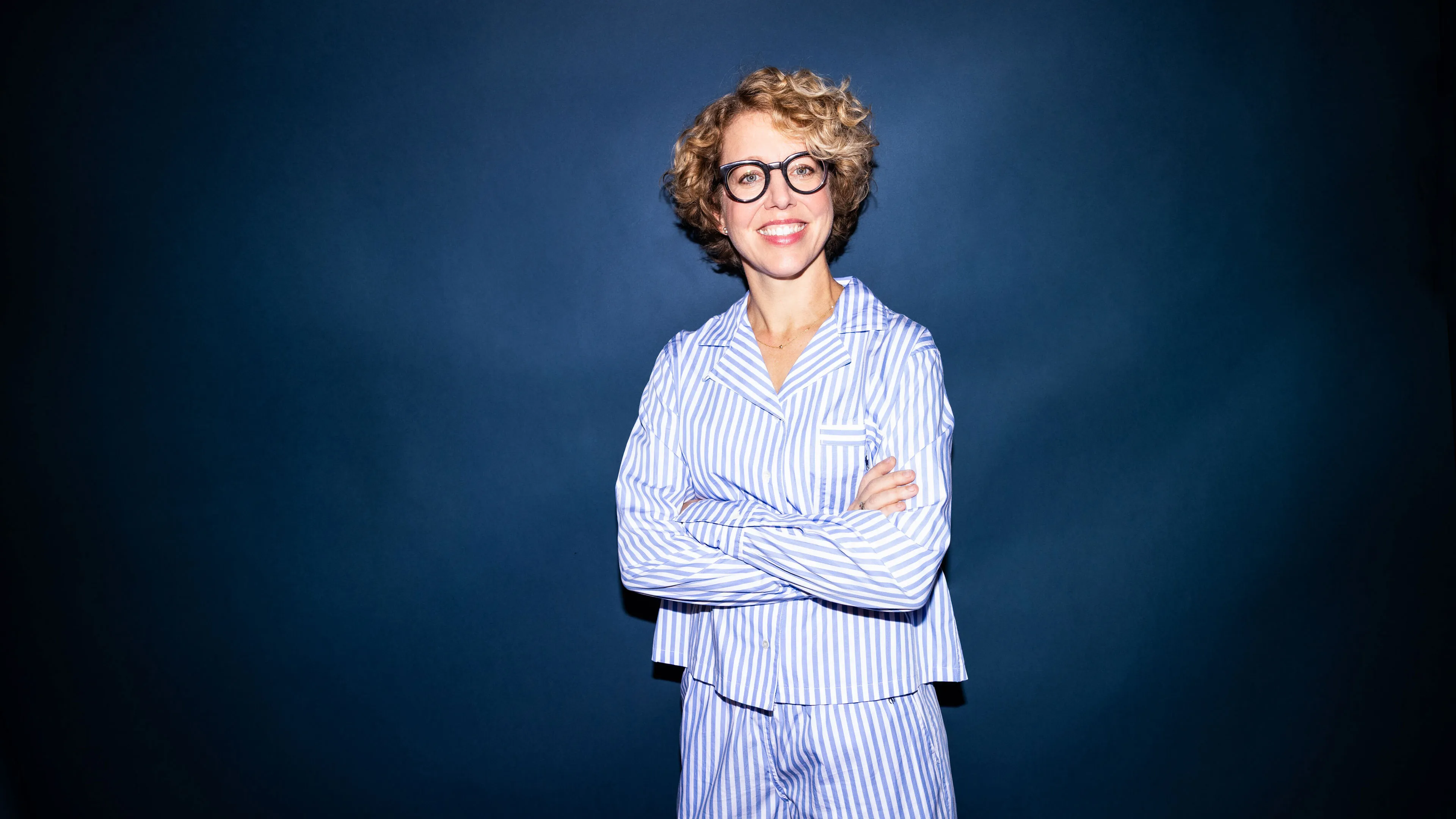 Lächelnde Frau mit lockigem blonden Haar und runder Brille, die blau-weiß gestreiften Pyjamaanzug trägt und vor einem dunklen Hintergrund steht.
