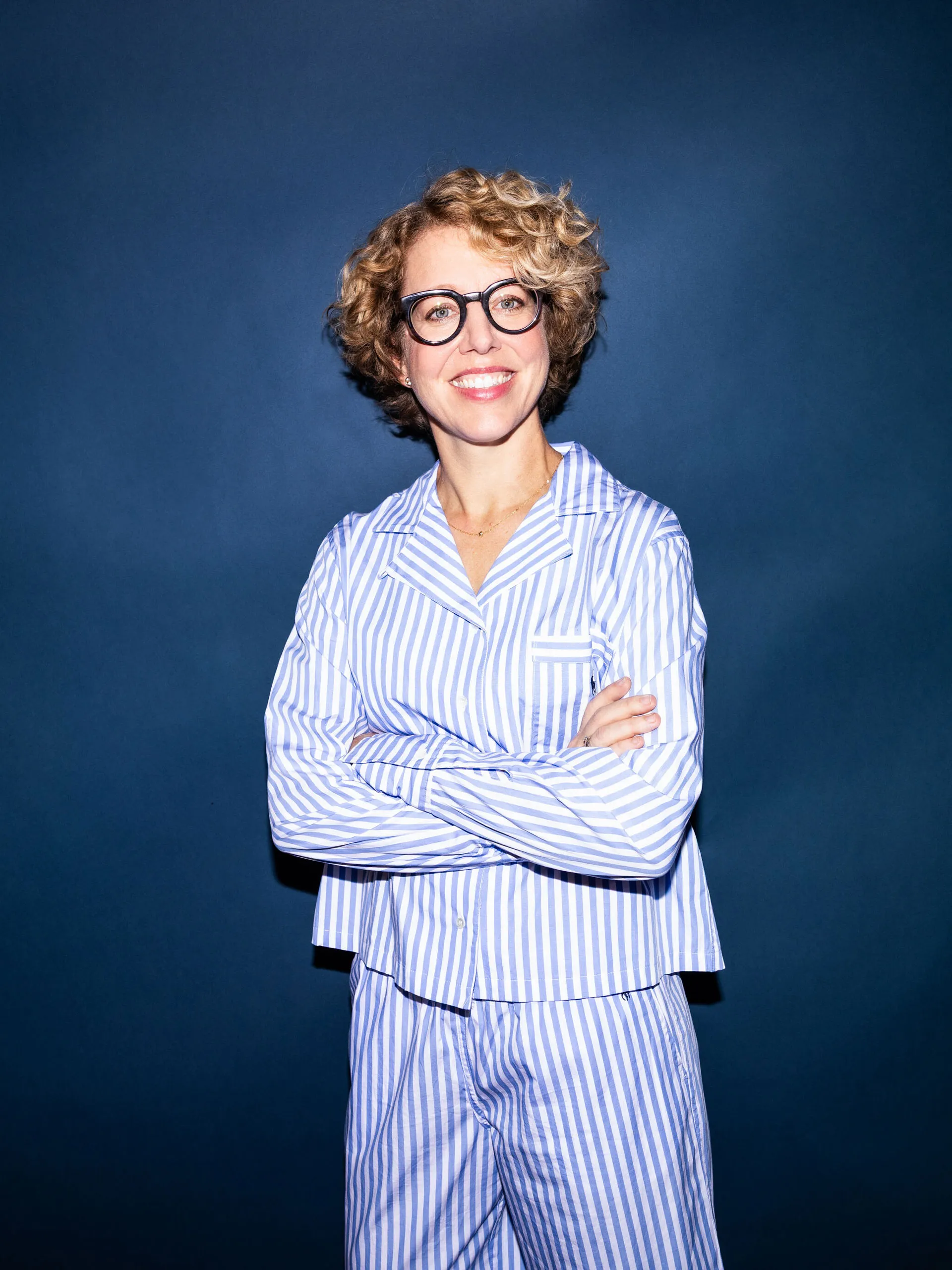 Frau mit kurzen lockigen blonden Haaren und Brille trägt blau-weiß gestreifte Pyjamahose und -hemd vor blauem Hintergrund.