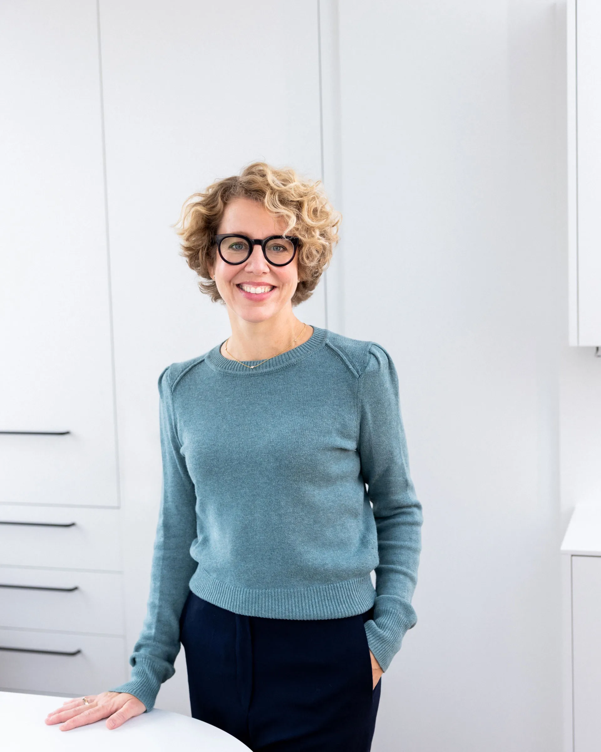 Frau mit lockigem blondem Haar, schwarzer Brille und blauem Pullover lächelt in einer hellen, weißen Küche.