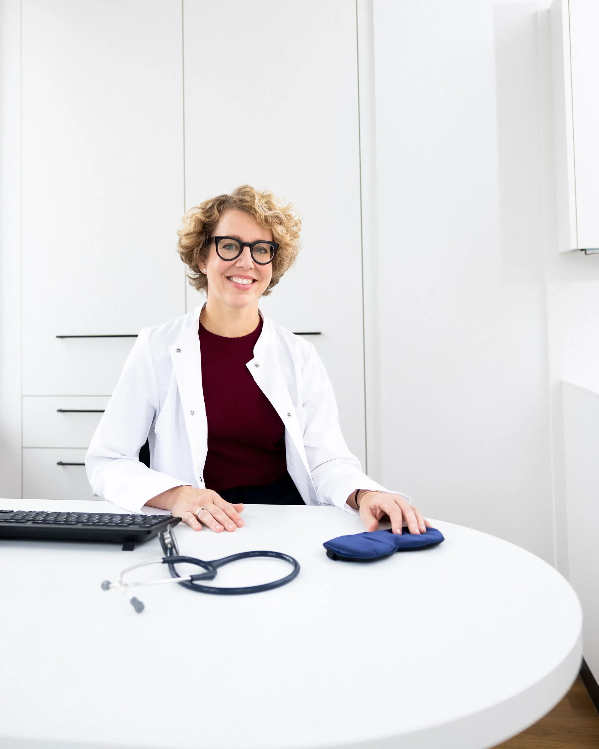 Lächelnde Ärztin mit weißen Kittel und Brille sitzt an einem weißen Schreibtisch mit Stethoskop und Augenmaske.