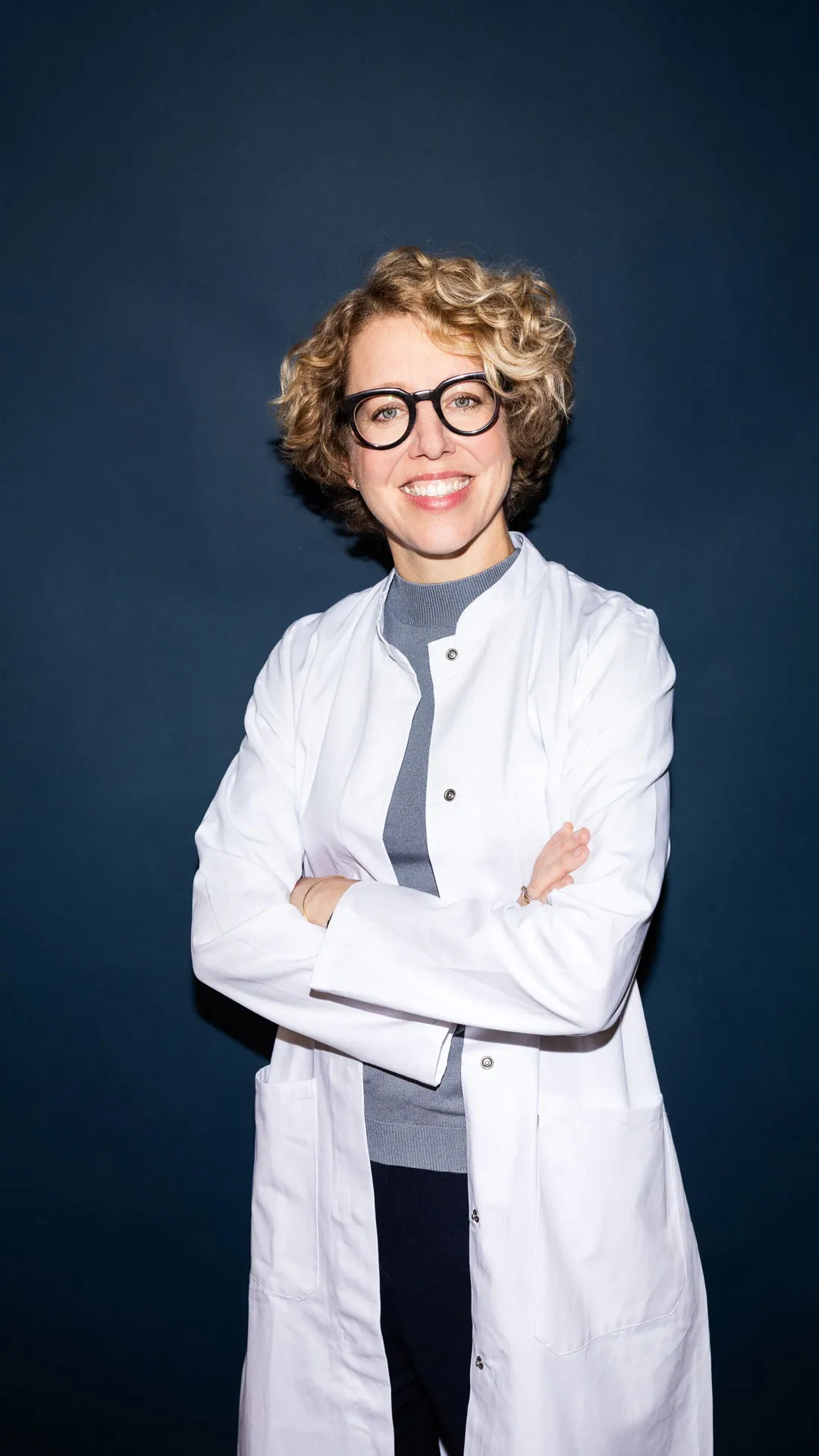 Lächelnde Frau mit lockigem Haar und Brille, trägt weißen Laborkittel und steht vor dunklem Hintergrund mit verschränkten Armen.
