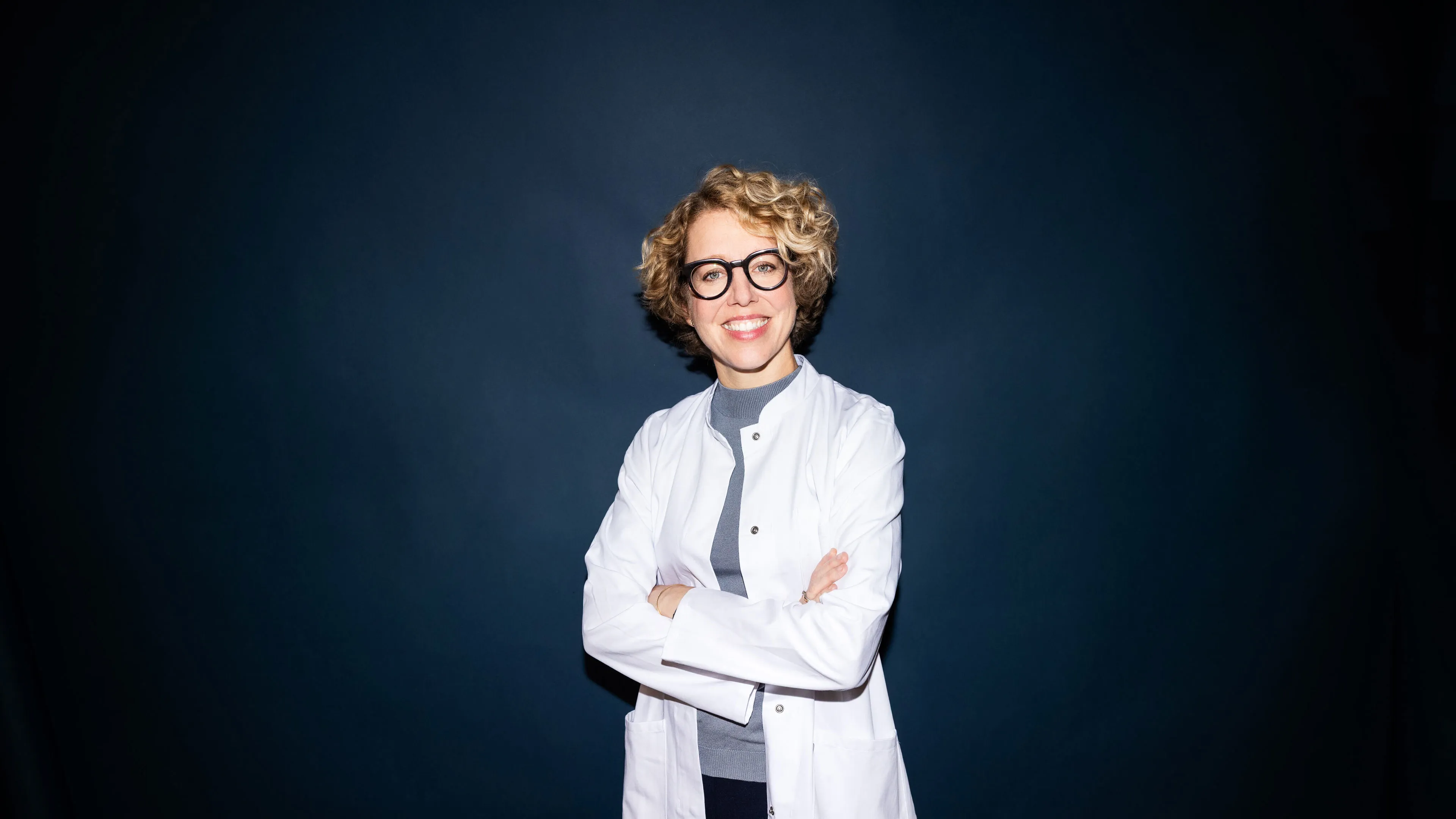 Lächelnde Frau mit lockigem blonden Haar, schwarzer Brille und weißem Arztkittel vor dunkelblauem Hintergrund.