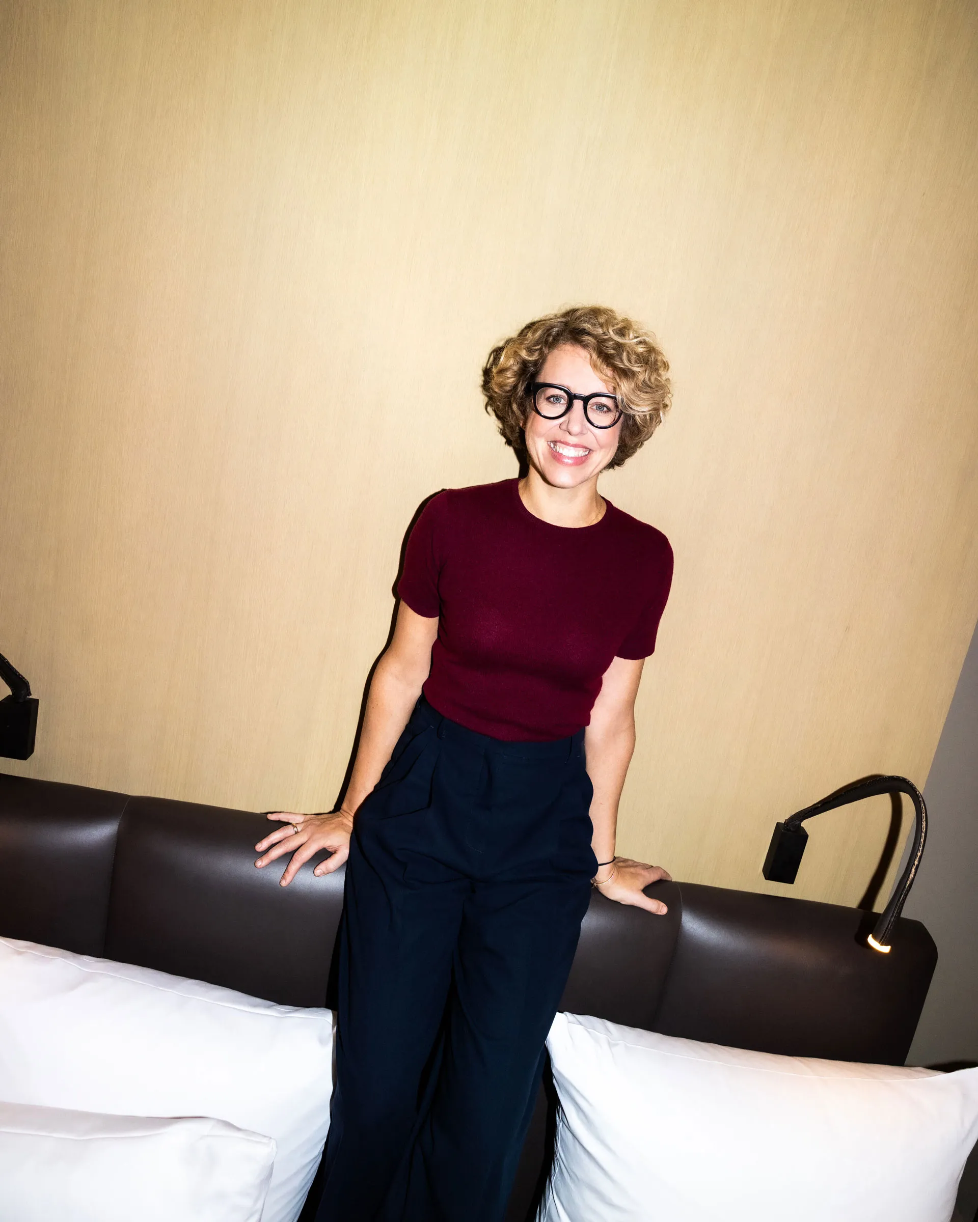 Frau mit lockigem blonden Haar, die eine rote Bluse und eine schwarze Hose trägt, sitzt lächelnd auf einem Sofa mit zwei weißen Kissen und beiger Wand im Hintergrund.