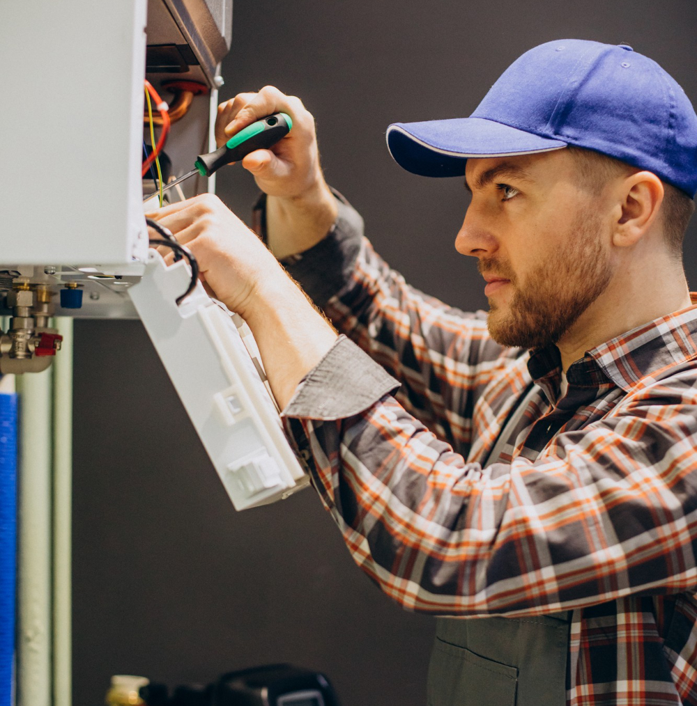 Man in a blue cap and plaid shirt using a screwdriver to repair a wall-mounted device.