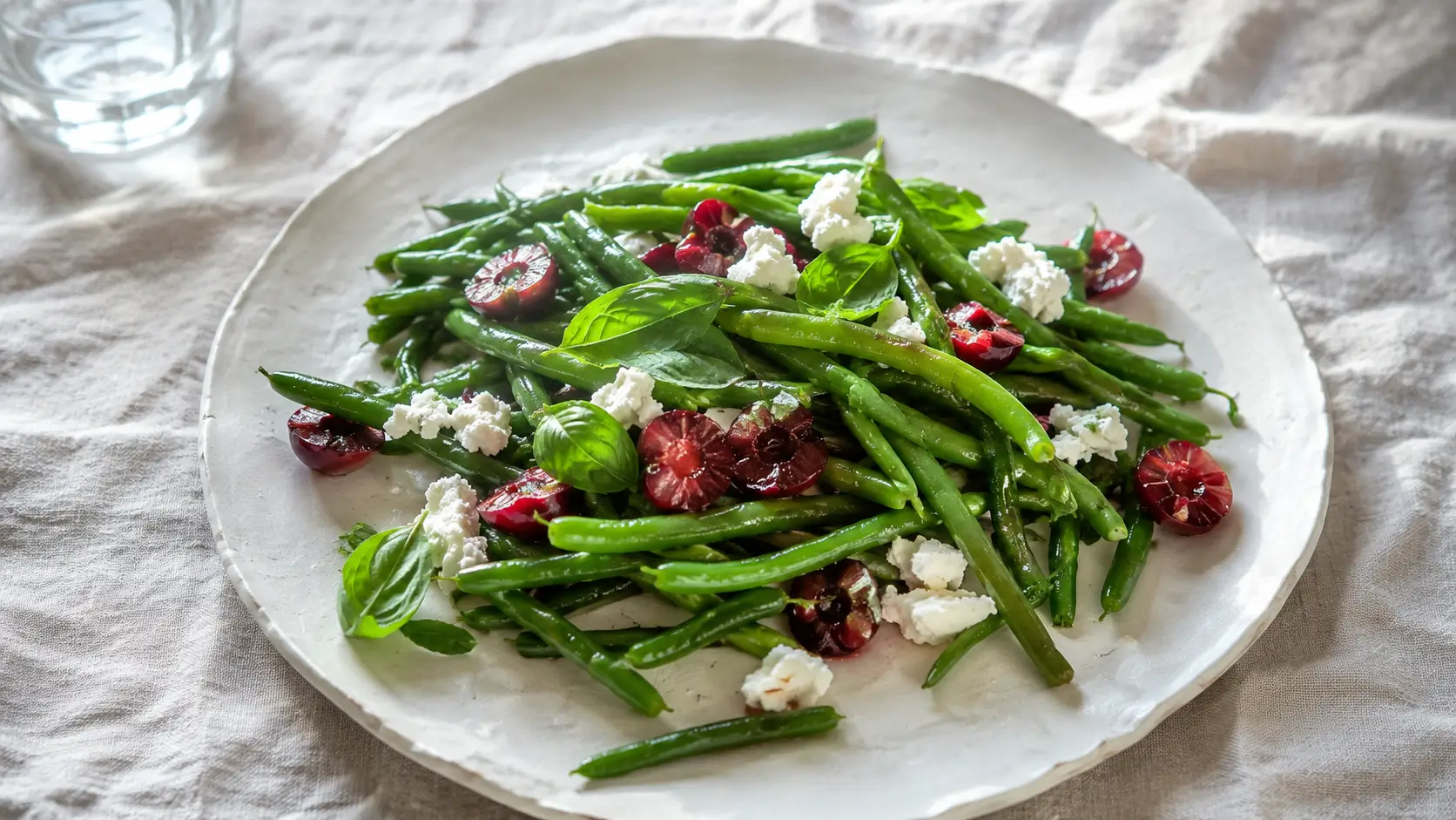 Salade tiède de haricots verts croquants et cerises rouges avec chèvre frais émietté et basilic, servie dans une assiette en céramique blanche
