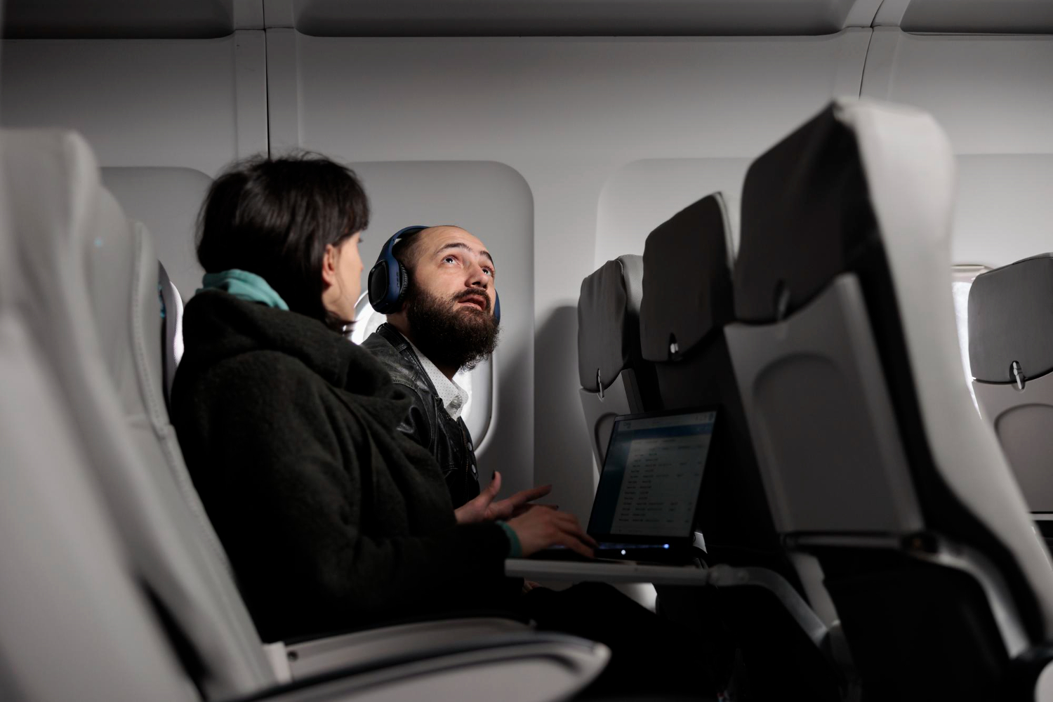 A man with a beard and a woman wearing headphones sit in a plane.