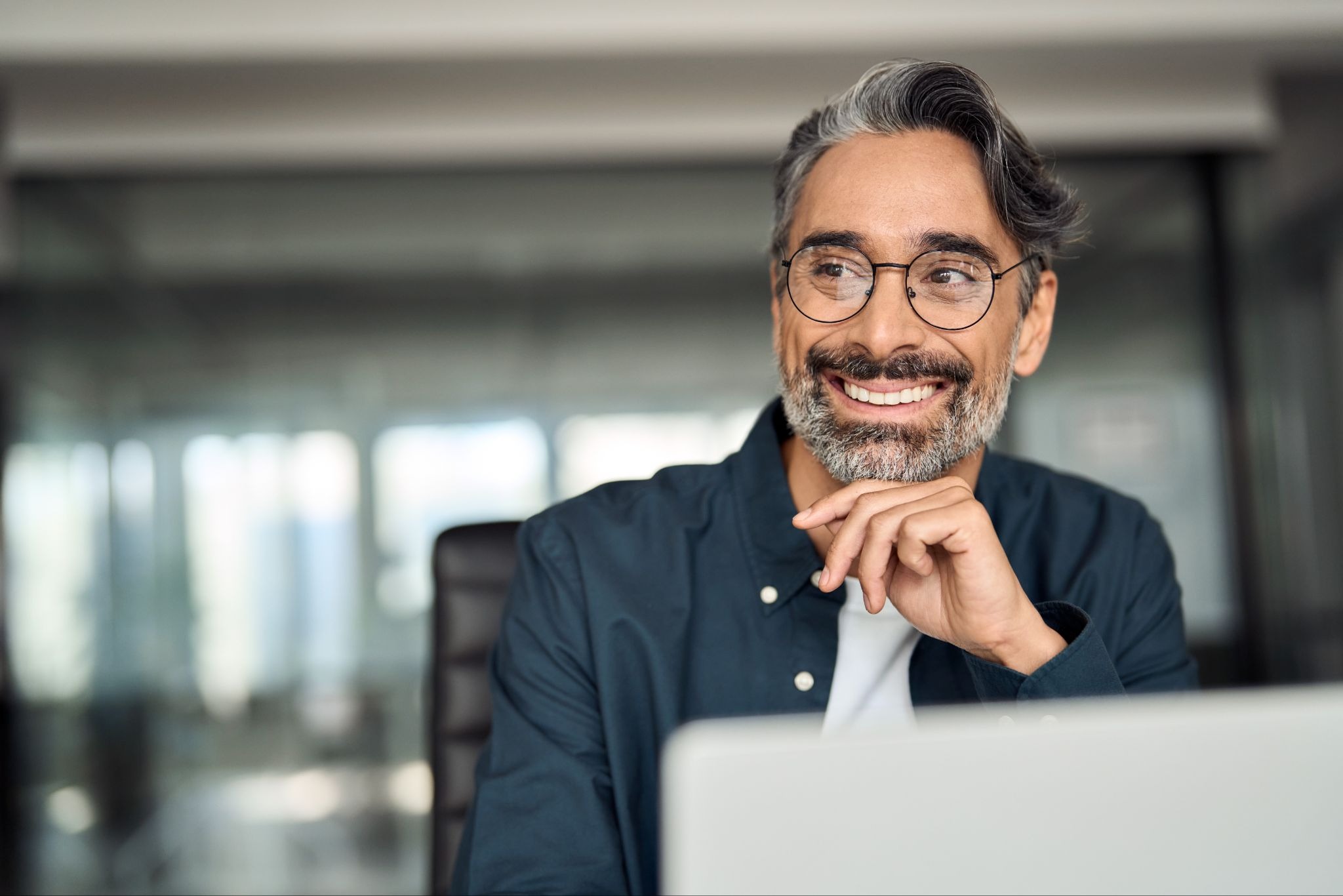 The man rests his chin on one hand and smiles warmly, suggesting he is engaged in a pleasant conversation, reflecting on an idea, or feeling confident about his work. A laptop is positioned in front of him, indicating he may be working remotely, attending a virtual meeting, or completing professional tasks.