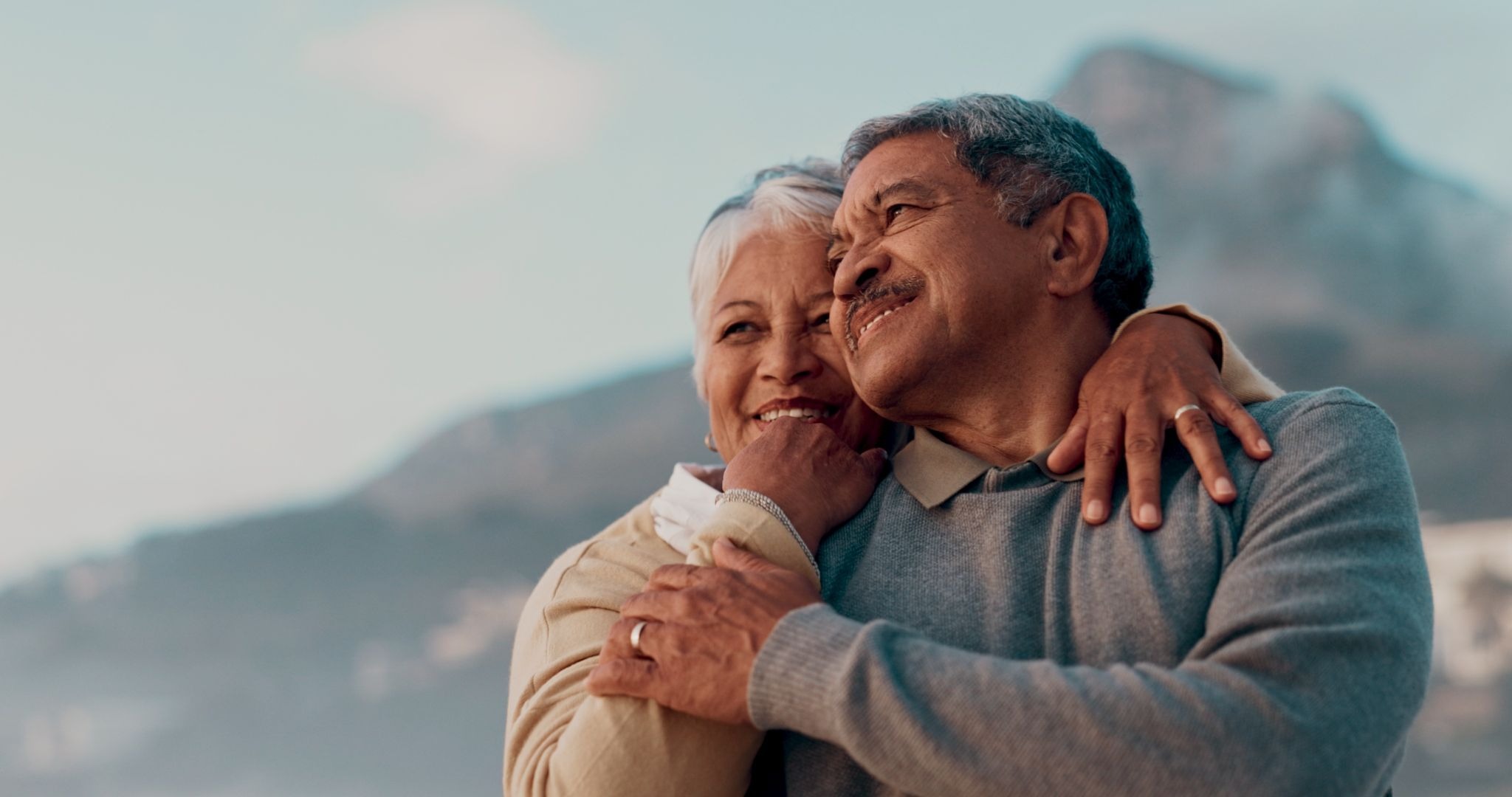 The man, who has short gray hair and a gray mustache, is wearing a gray long-sleeve sweater. He smiles gently while looking toward the distance, conveying contentment and happiness. The woman, with short white hair, stands beside him with one arm wrapped around his shoulders and the other resting on his chest. She smiles affectionately as she looks at him, highlighting a close and loving relationship.