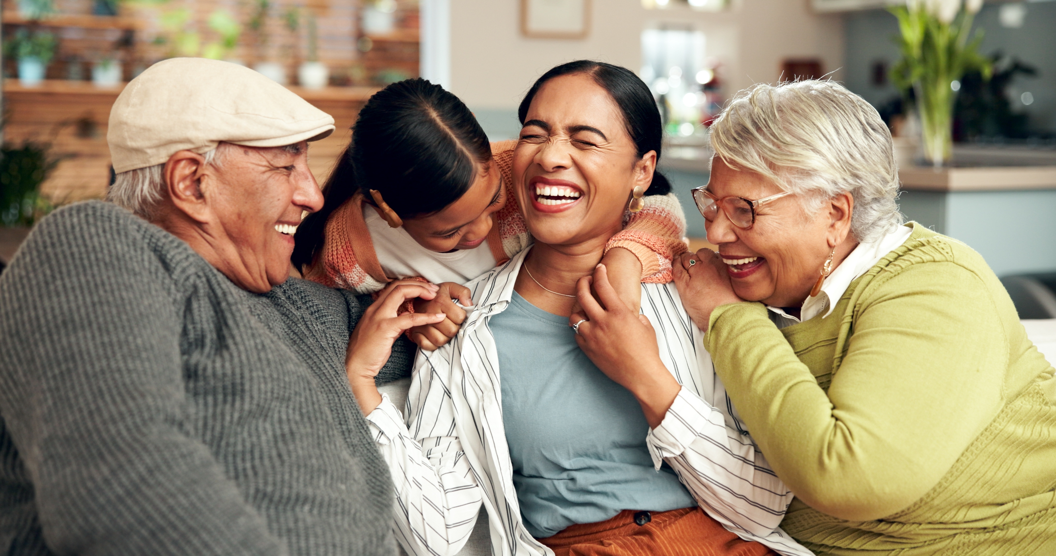 An older couple is sharing a happy moment with a younger woman and a child, all gathered closely together on a couch. The young girl is hugging the woman from behind while both older adults lean in with smiles, creating a sense of affection and togetherness. Everyone appears to be laughing, suggesting a candid and heartfelt family interaction.