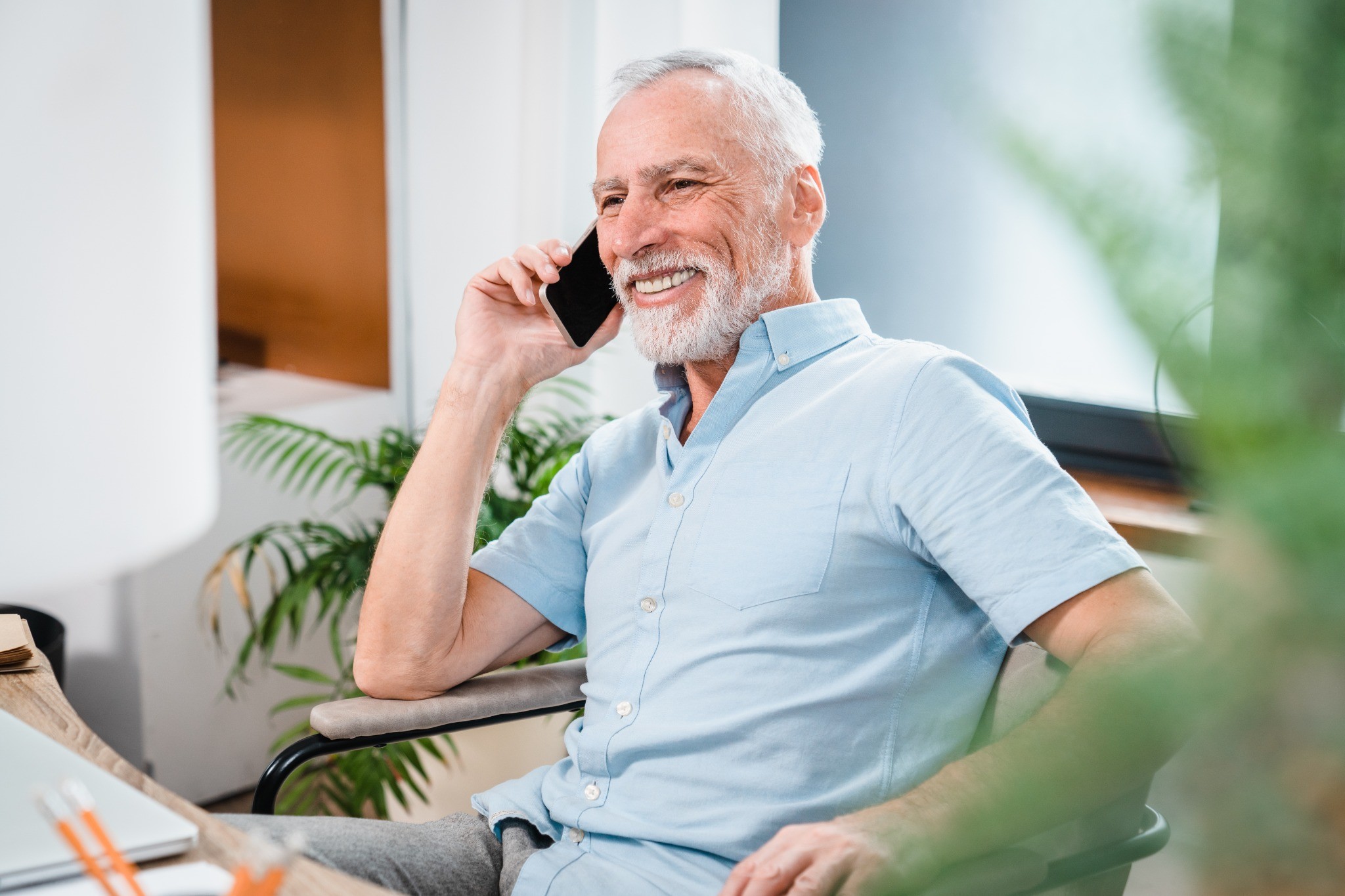 A smiling older man with gray hair and a neatly trimmed white beard is sitting comfortably in a bright, modern home while talking on his smartphone. He is wearing a light blue short-sleeve button-up shirt and appears relaxed and happy during the conversation. The room is filled with natural light, with green indoor plants and contemporary furnishings creating a warm, inviting atmosphere. A laptop and notebook are visible on the desk in front of him, suggesting he may be discussing important personal or financial matters from home.