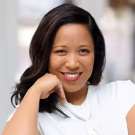 Smiling woman with long dark hair resting her chin on her hand against a blurred background.