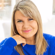 Smiling woman with blonde hair wearing a blue sweater, resting her chin on her hand.