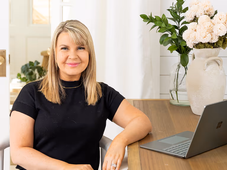 Smiling woman with blonde hair sitting at a wooden desk with a laptop and white flowers in a vase.