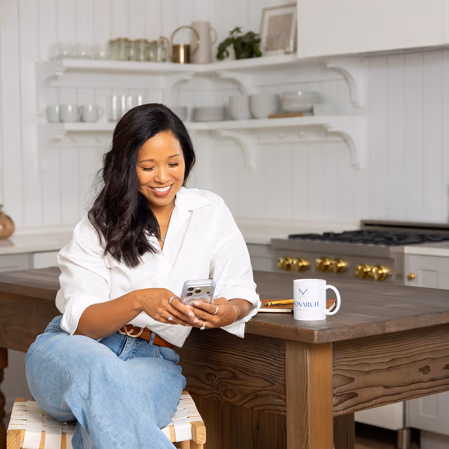 Smiling woman with black hair wearing a white shirt and jeans, sitting on a stool at a wooden kitchen island, holding a smartphone with a mug on the counter beside her.