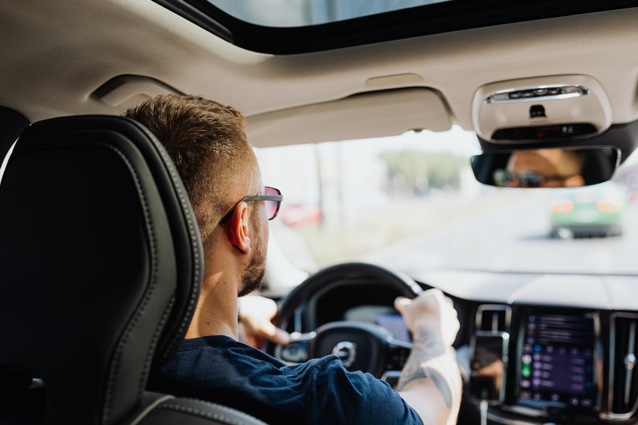 Man with glasses driving a modern car, seen from the back seat focusing on the steering wheel and dashboard.