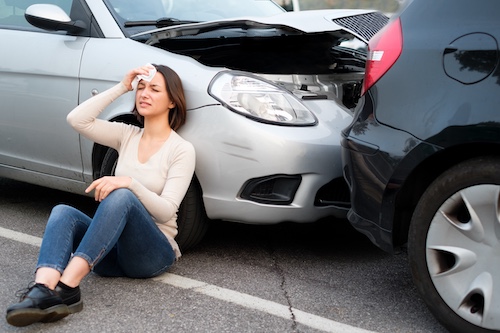 Woman sitting on the ground beside a silver car with a damaged front after a rear-end collision with a black car.
