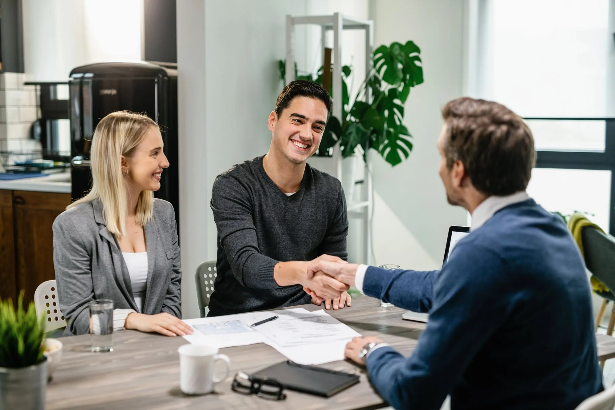 A smiling young man shakes hands with an advisor across a table, while a woman sits beside him, both looking happy highlighting the welcoming, client-focused approach of an independent insurance agency in South Florida.