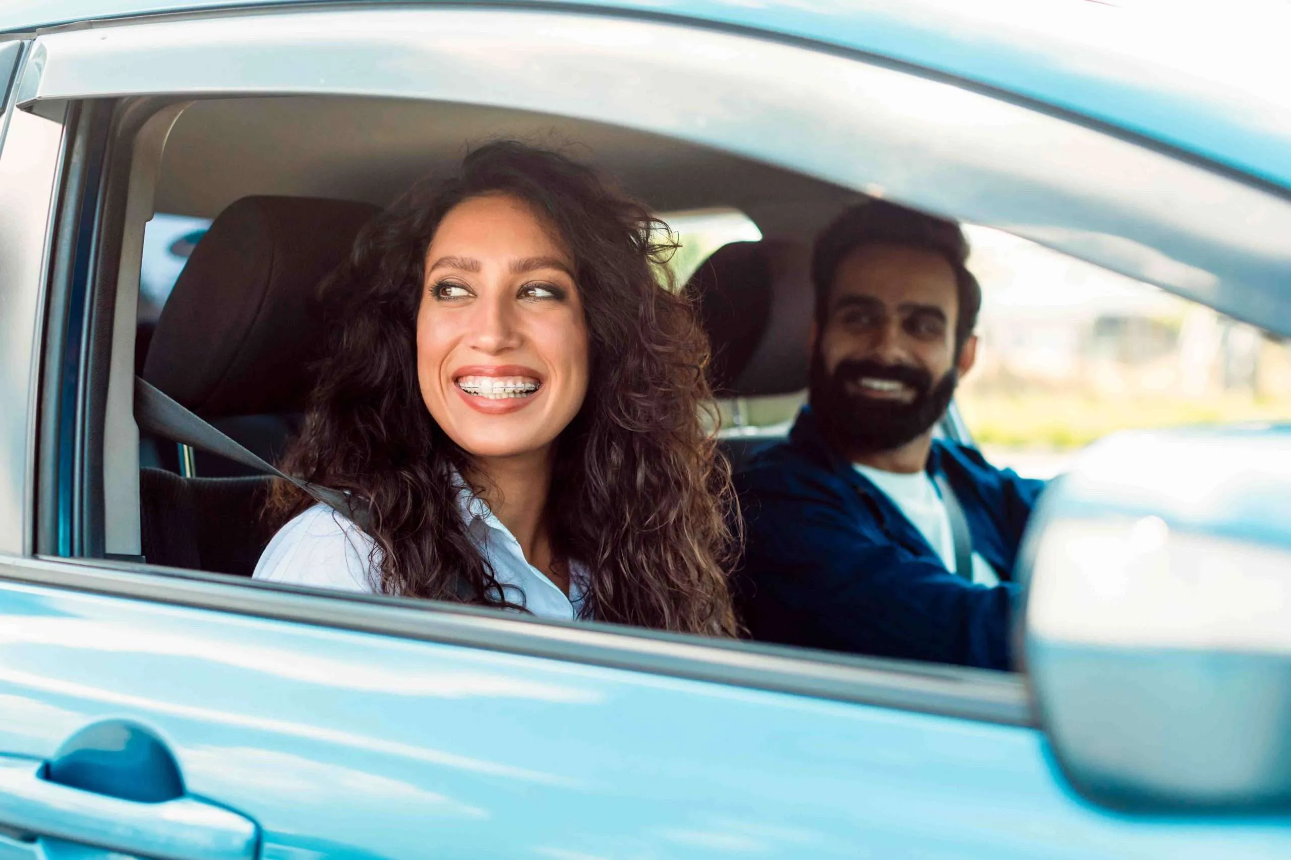 Smiling woman with braces sitting in the passenger seat of a car wearing a seatbelt, and a man driving and looking at her.