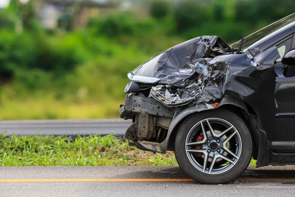 Black car with severe front-end damage parked by roadside with green grassy area in background.