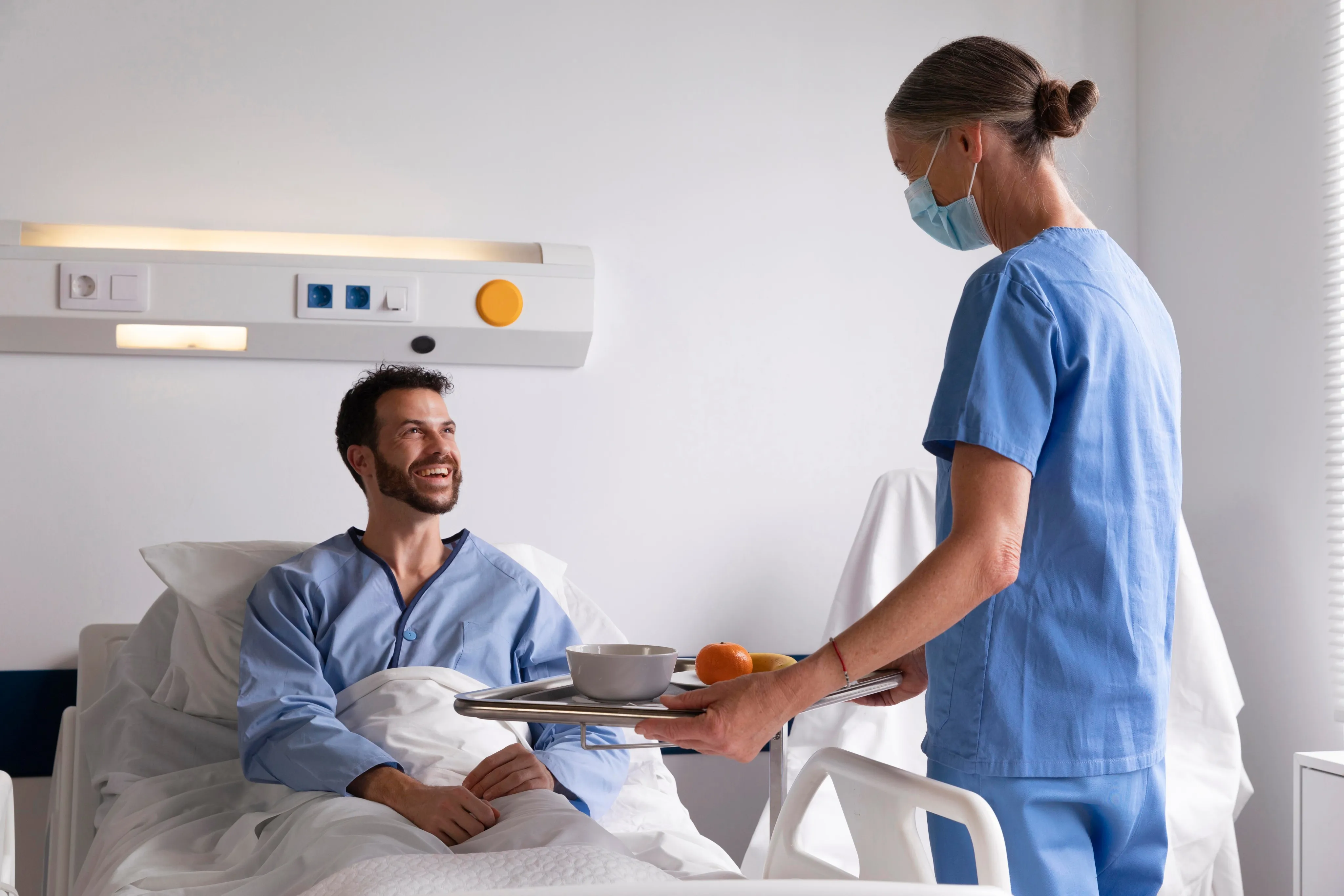Nurse wearing a mask serving food tray to a smiling male patient in a hospital bed.