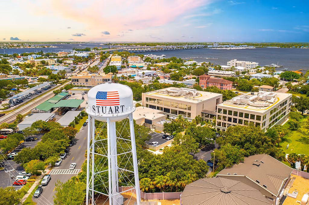 Aerial view of Stuart, Florida, showing a water tower with an American flag and the word 'STUART,' nearby buildings, roads, and the waterway with a bridge in the background during sunset.