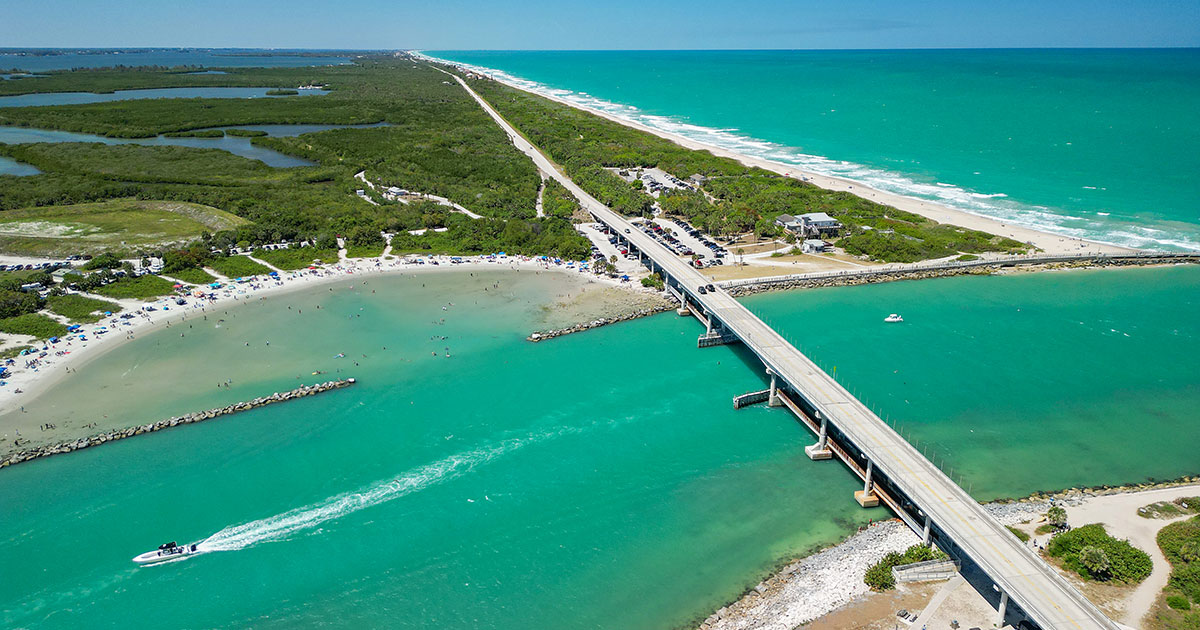 Aerial view of a coastal bridge over turquoise water with a boat leaving a white trail and a beach with people near green vegetation.