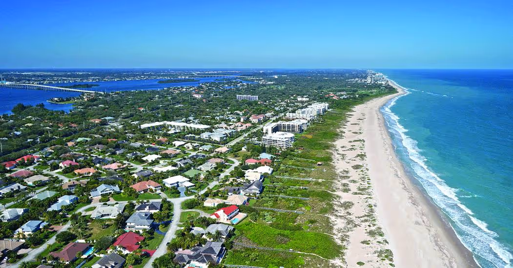 Aerial view of Vero Beach, Florida, featuring a long stretch of quiet sandy shoreline along the Atlantic Ocean. Residential neighborhoods with tree-lined streets and coastal homes sit just inland, while mid-rise condominiums overlook the beach. The Indian River Lagoon and a bridge crossing the waterway are visible in the distance, highlighting Vero Beach’s scenic blend of oceanfront and riverfront landscapes