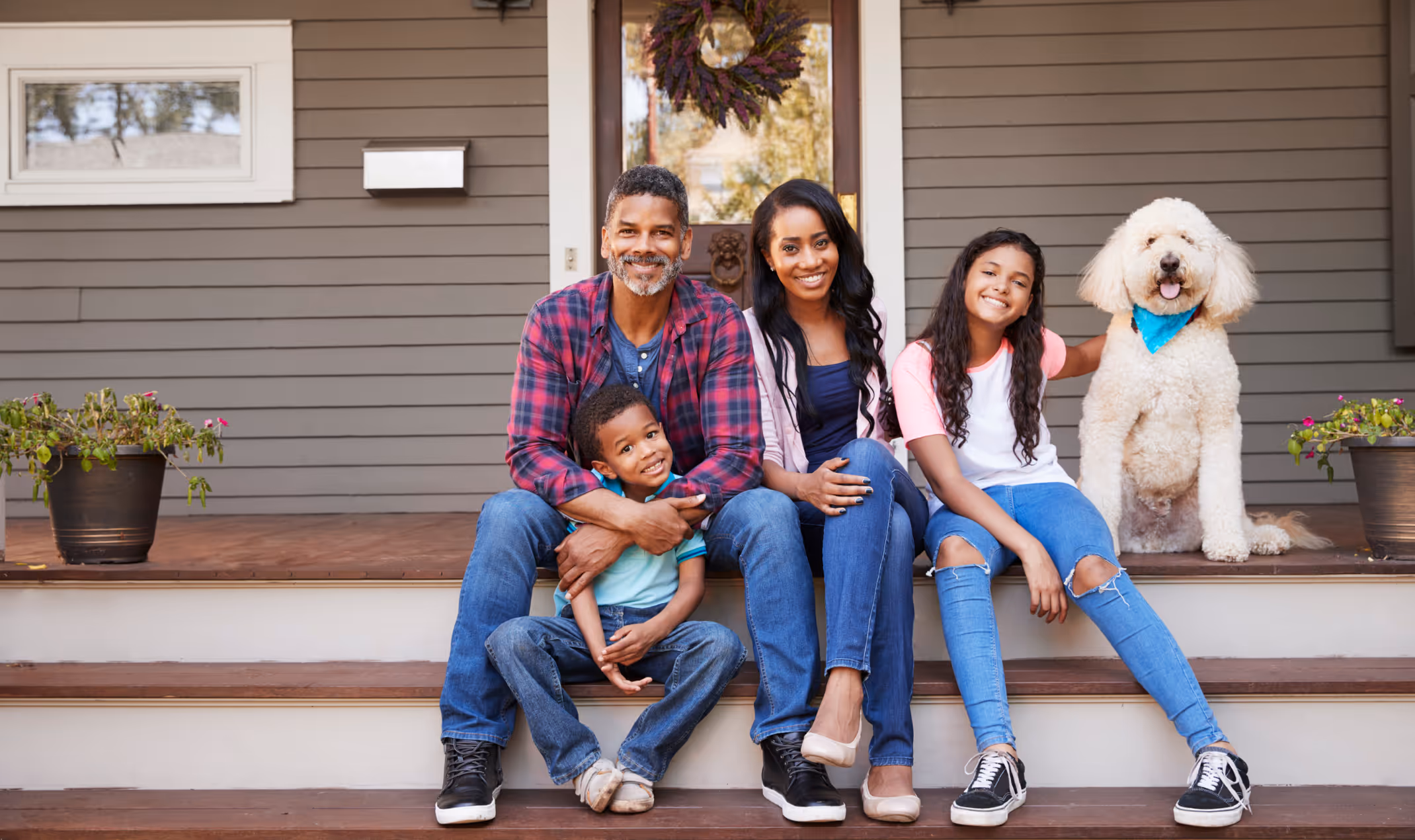 Smiling family of four sitting on wooden front porch steps with a large white dog wearing a blue bandana.