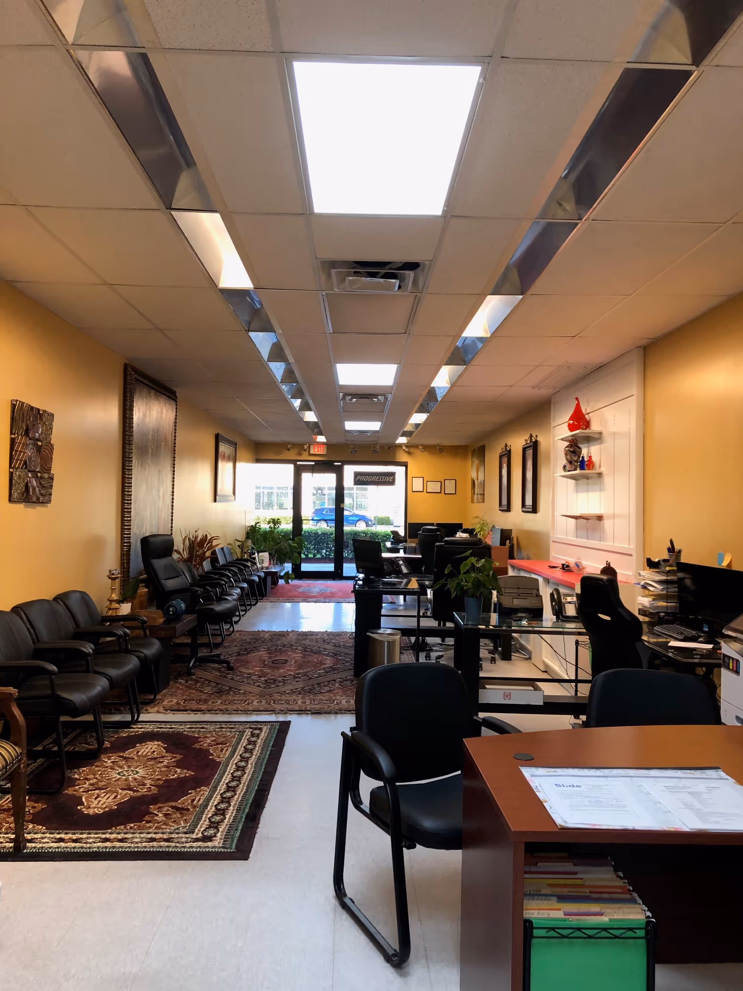 Office interior with black chairs lined up along a yellow wall, desks with computers, plants, and decorative rugs on a white floor.