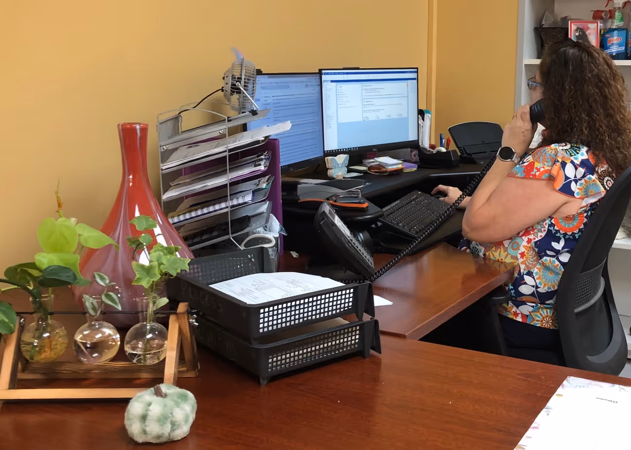 Woman with curly hair talking on a desk phone in a colorful floral shirt, seated at a desk with dual monitors, paper trays, and small plants.