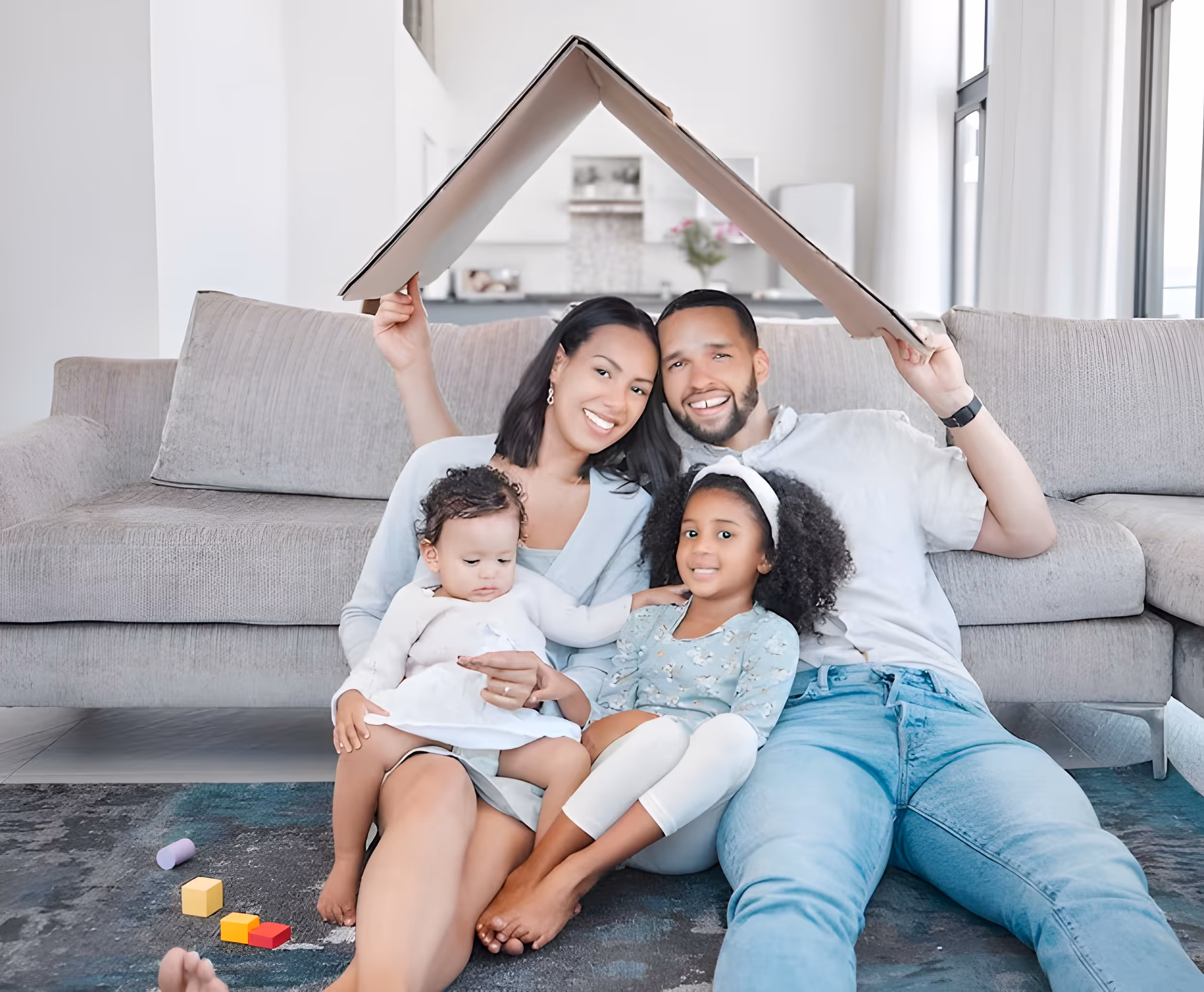 Smiling family of four sitting on the floor in their living room with parents holding a cardboard roof above them.