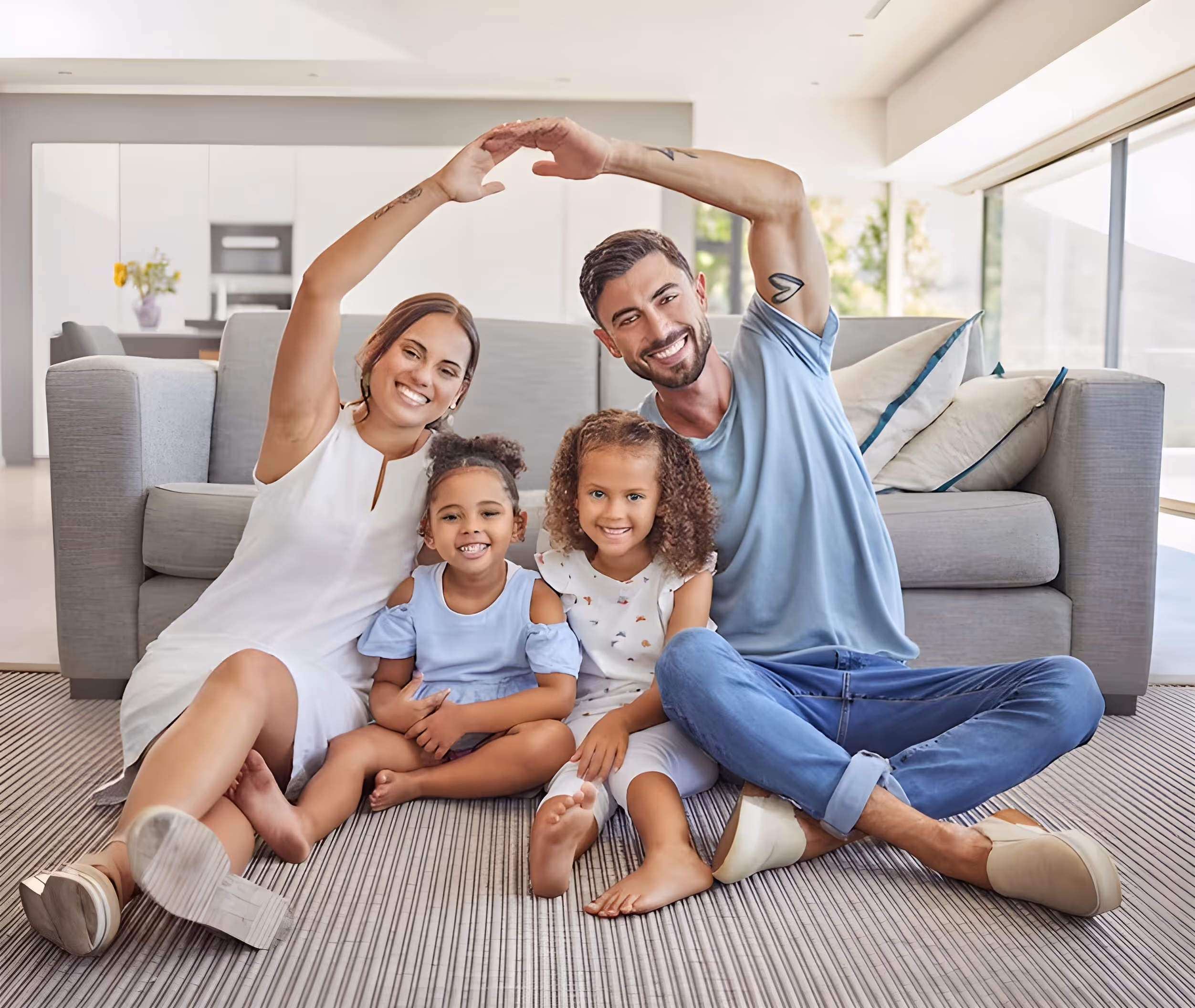 Smiling family of four sitting on a rug in a living room with father and mother forming a heart shape with their arms.